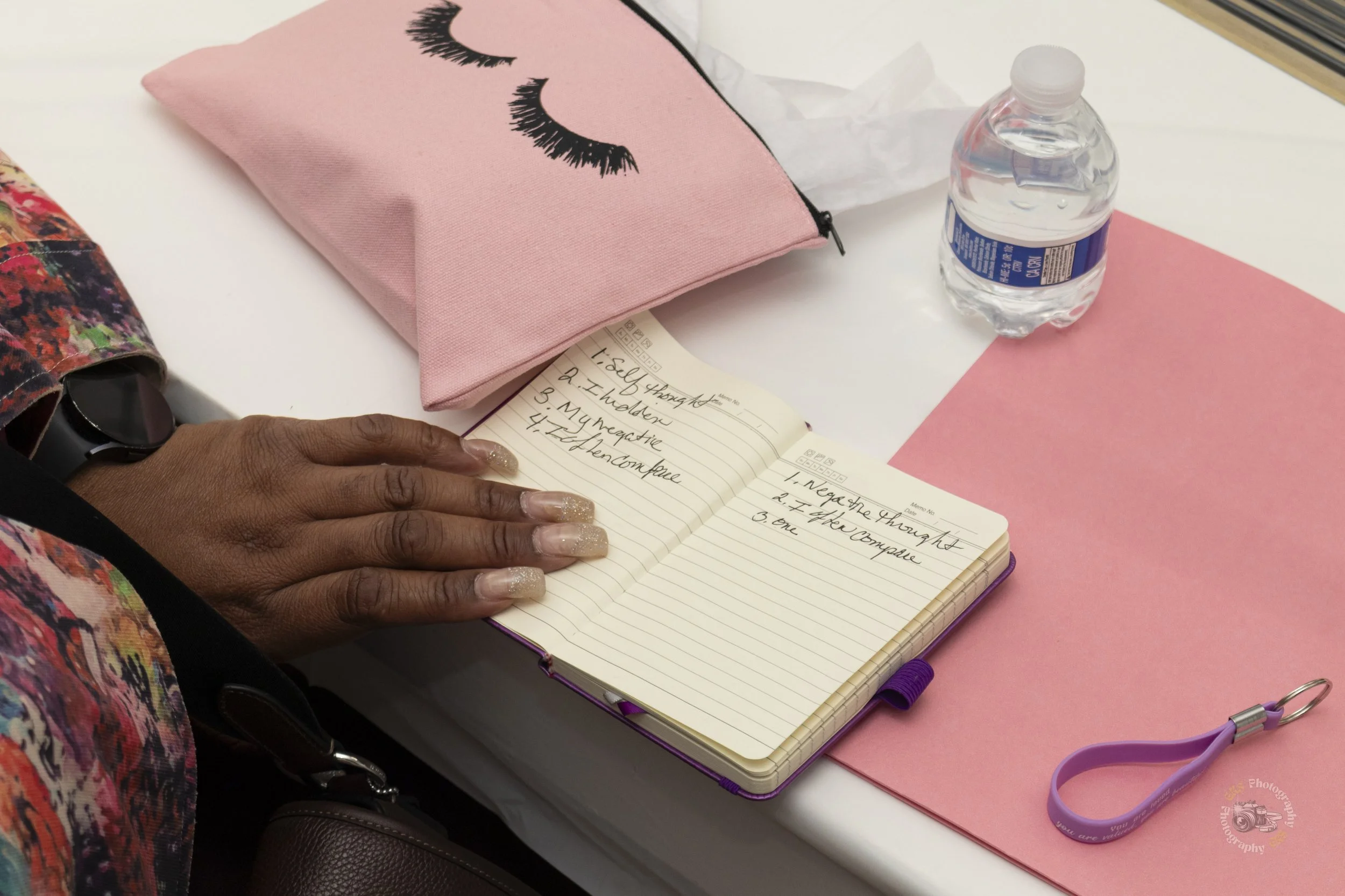 A person with painted nails writing in a notebook at a pink table, with a pink sleep mask, water bottle, and pink paper nearby.