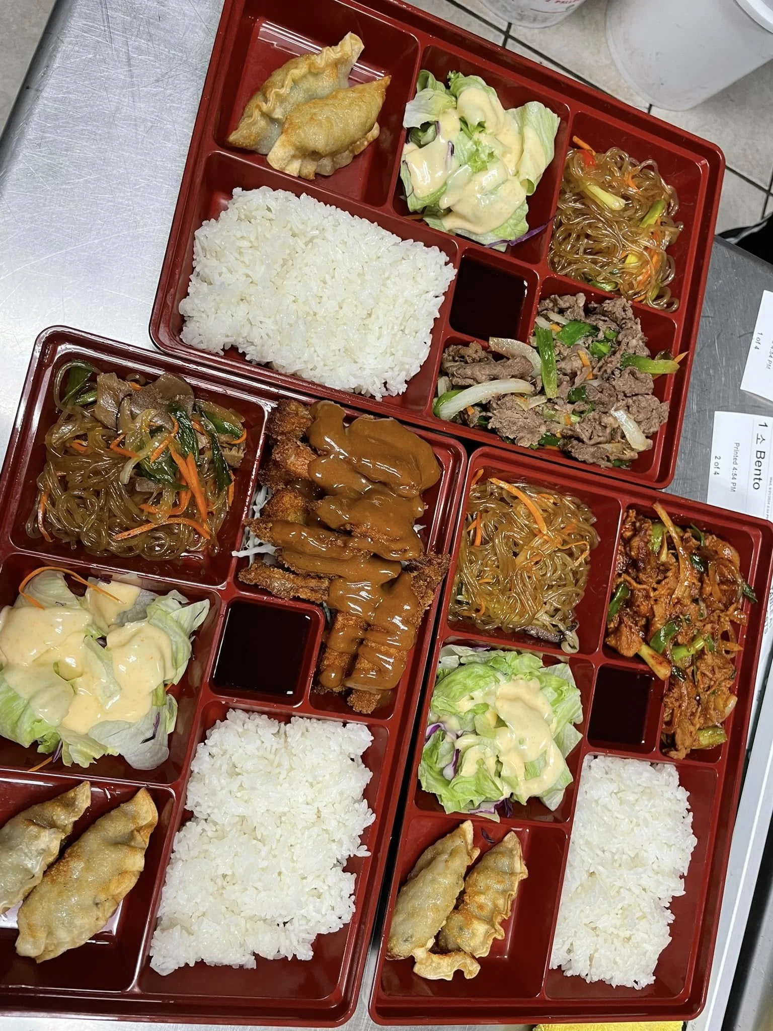Red bento boxes with rice, fried dumplings, salads, and Asian noodles placed on a metal table.