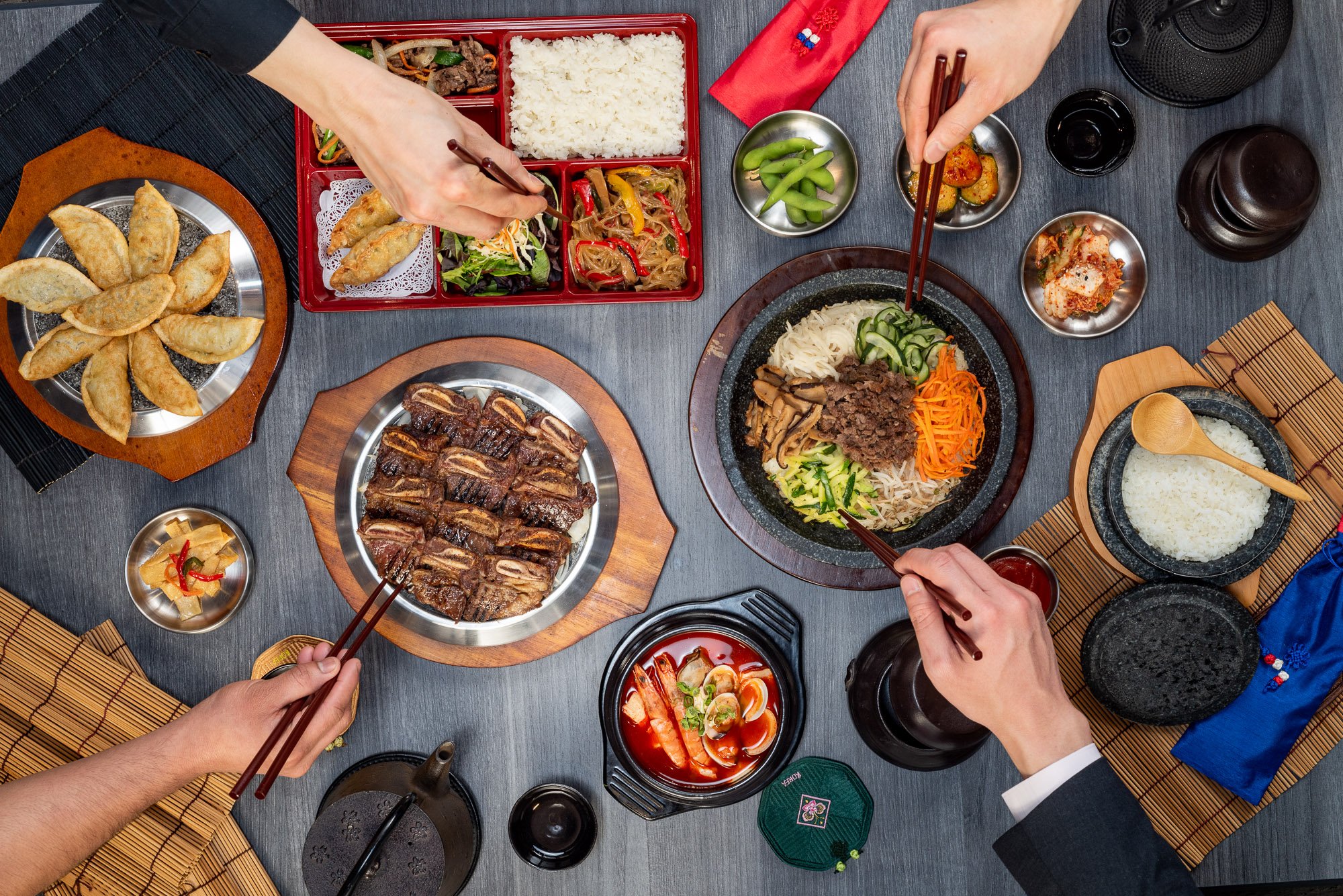 Top-down view of a Korean-style meal with various dishes including grilled meats, rice, kimchi, vegetables, and side dishes on a table with chopsticks and a teapot.