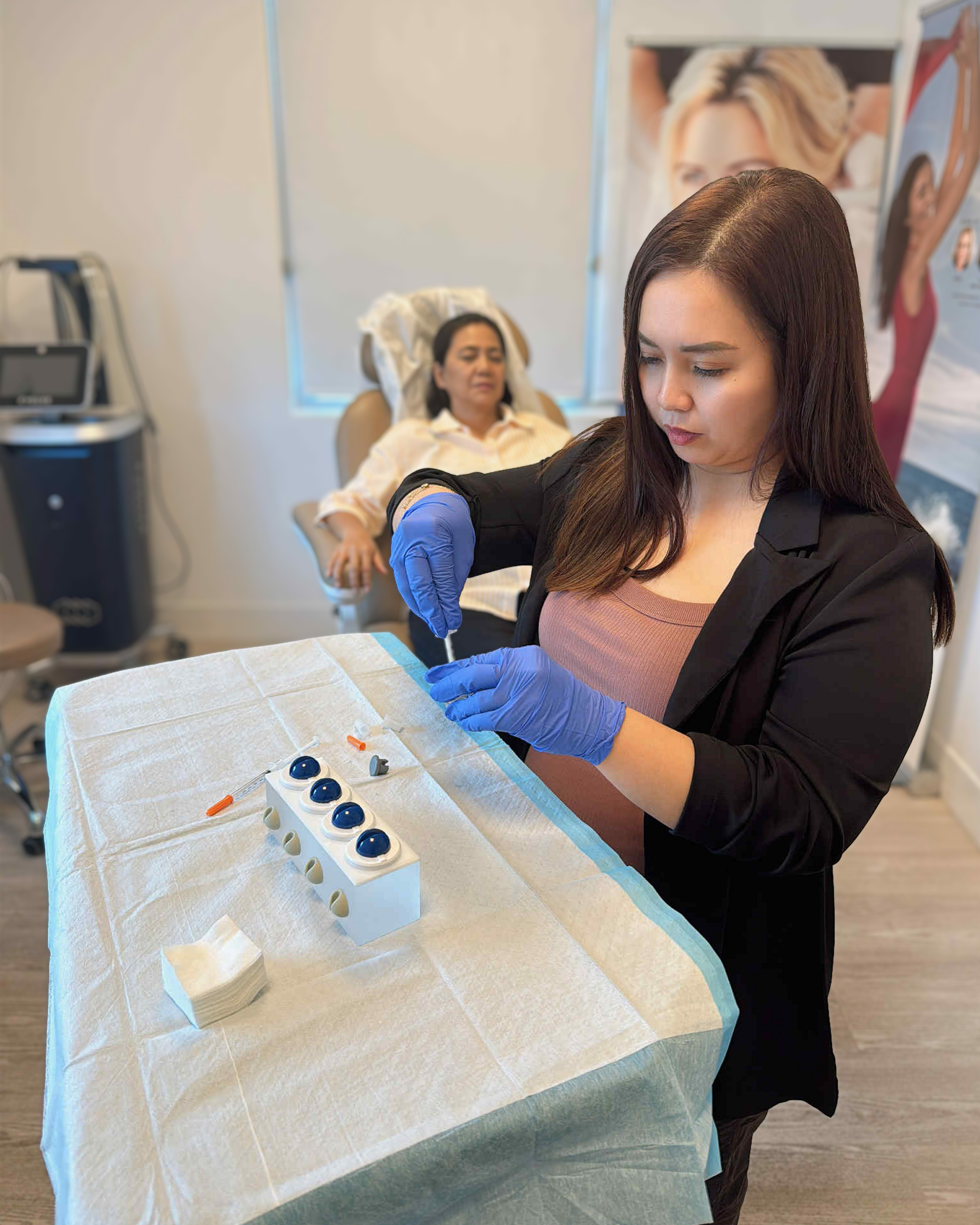 Injector in blue gloves preparing a syringe at the treatment tray while a client relaxes at Glow in Grace Aesthetics.