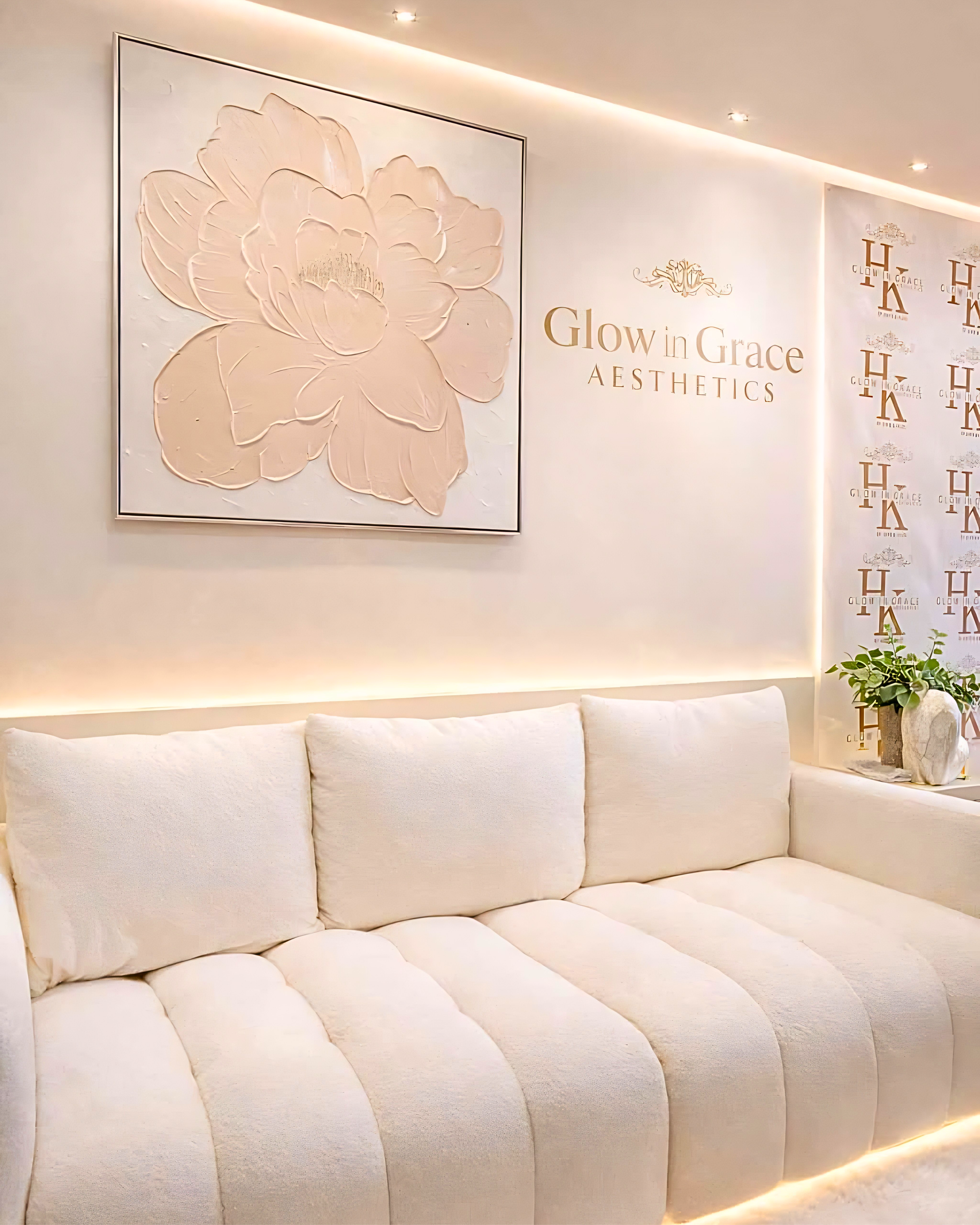 A white tufted sofa in a neutral-toned room with soft lighting, a large framed floral artwork on the wall, and decorative text that reads 'Glow in Grace Aesthetics' along with a logo, visible in the background.