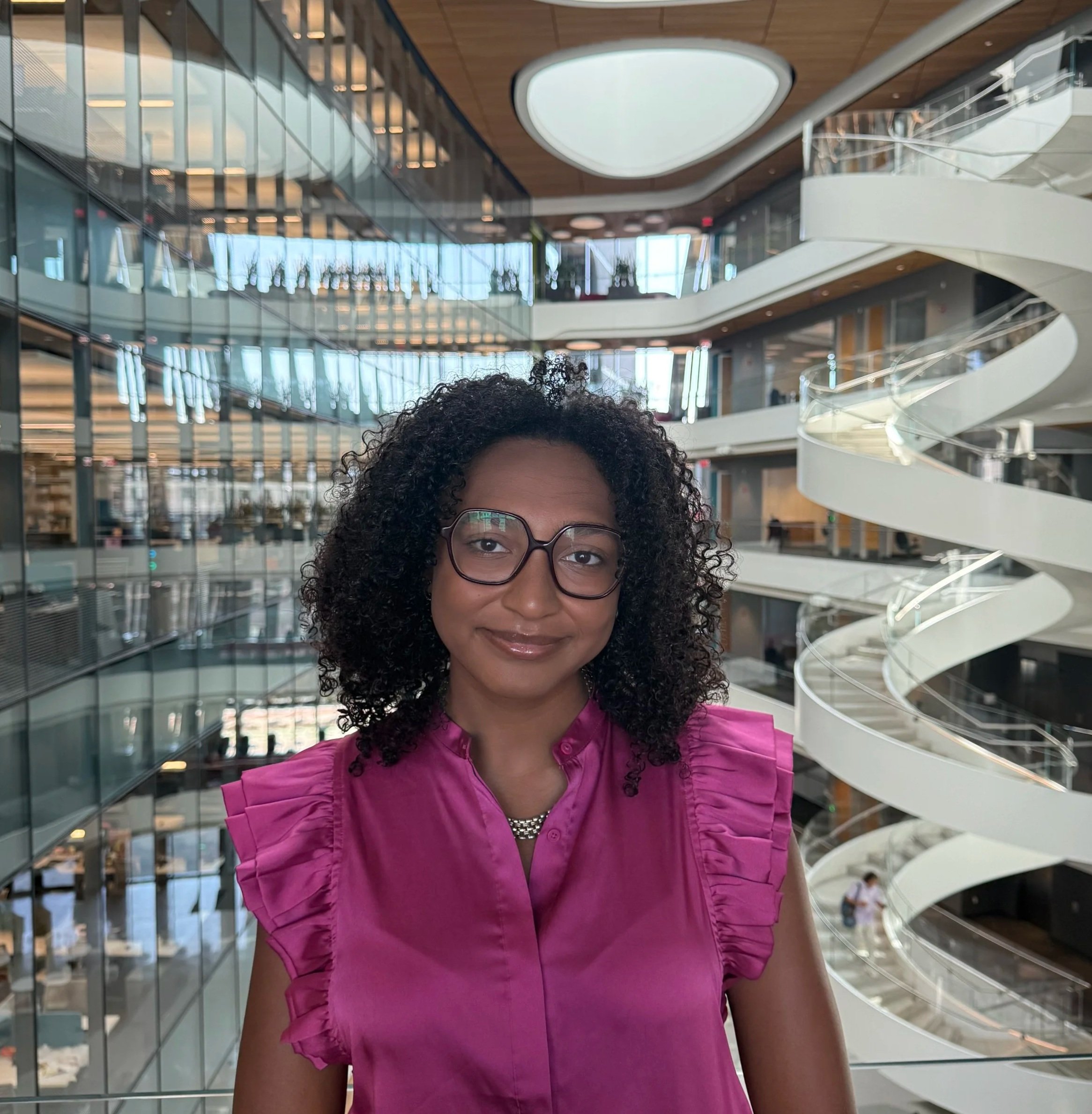 A woman with curly hair and glasses, wearing a pink top, standing inside a modern building with glass and spiral staircase architecture.