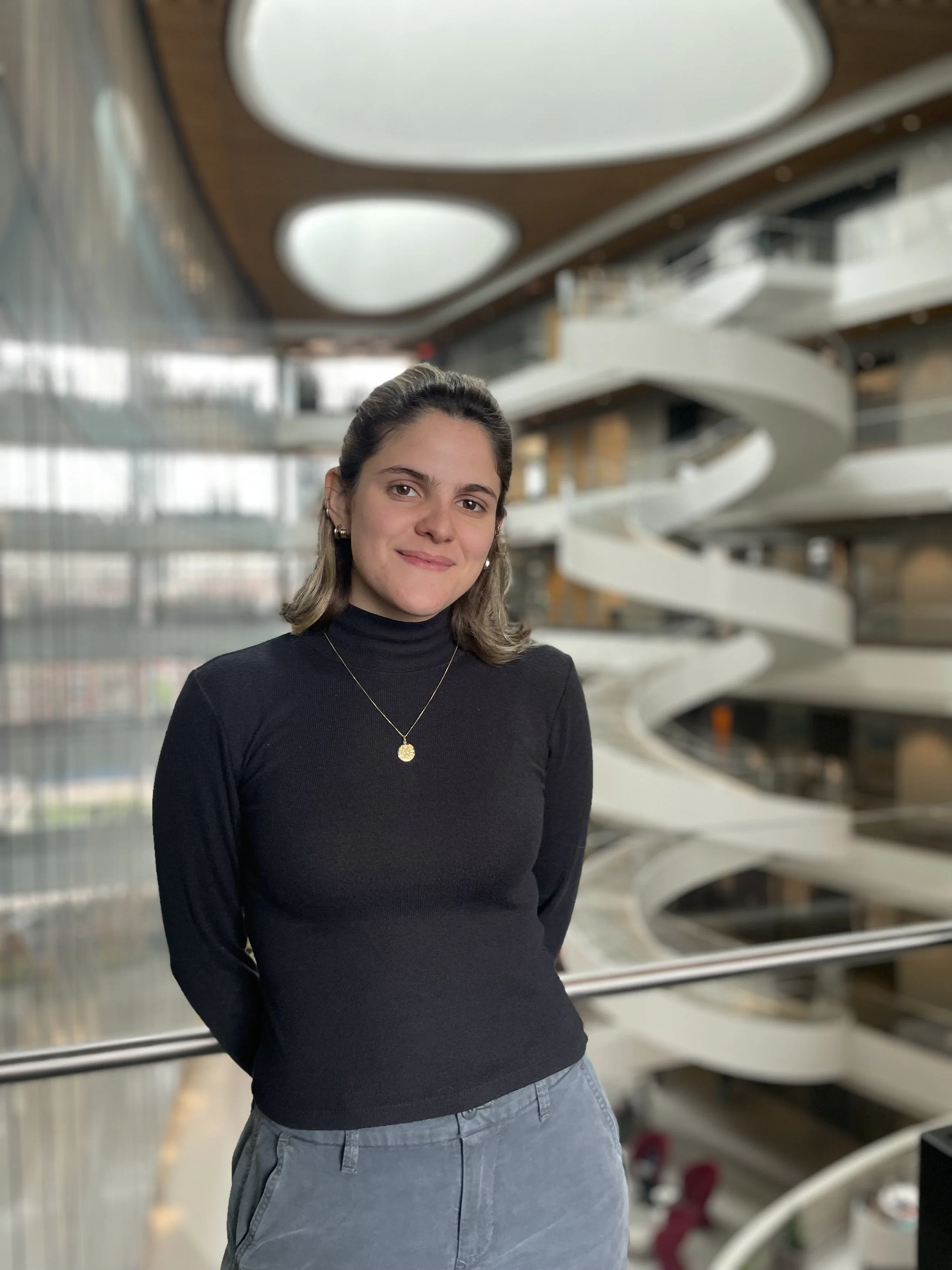 A woman with shoulder-length brown hair, wearing a black turtleneck and a gold necklace, stands inside a modern building with a spiral staircase and large circular ceiling lights in the background.