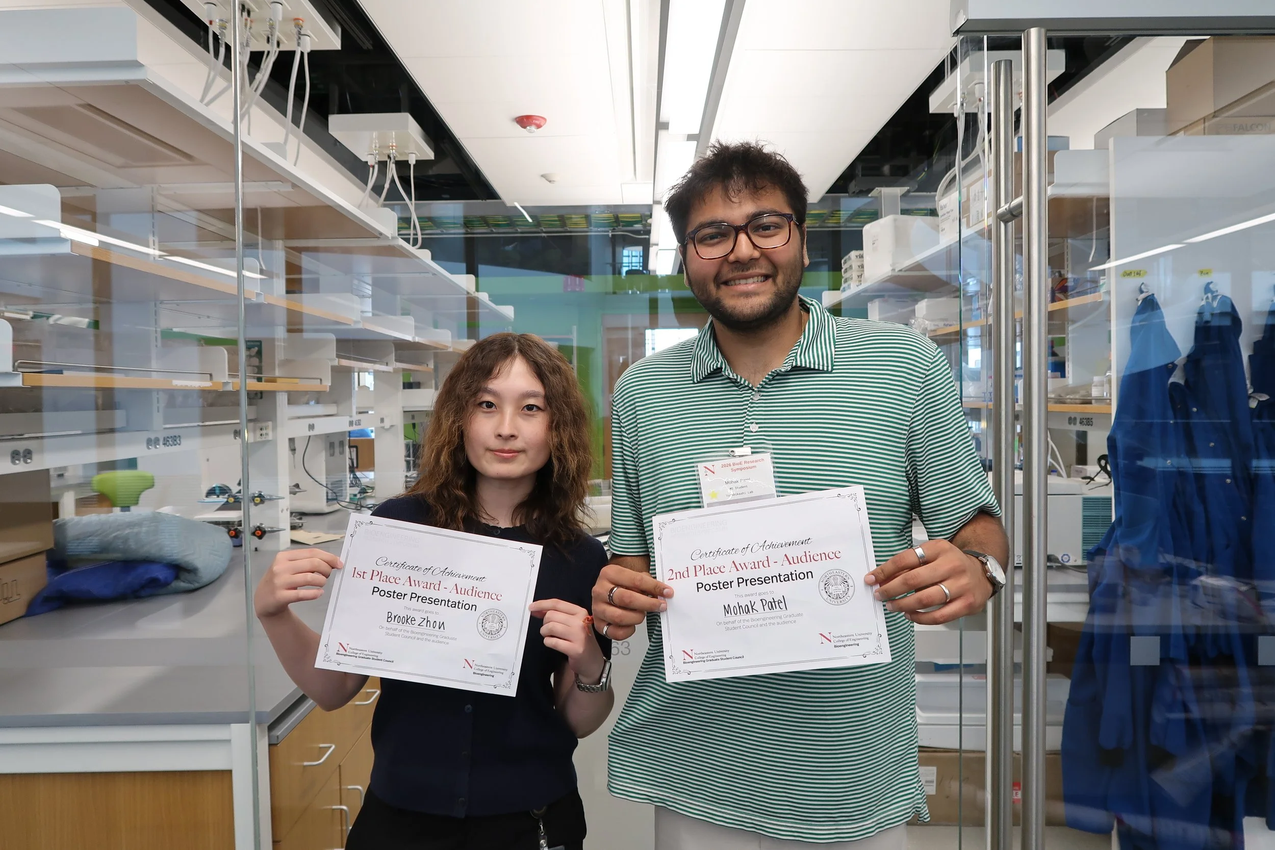 Two individuals holding certificates of achievement in a laboratory setting. The person on the left, a young woman with curly hair, is holding a certificate for 1st place in a poster presentation. The person on the right, a man with glasses and dark hair, is holding a certificate for 2nd place in a poster presentation. They are standing in front of laboratory equipment and glass display cases.