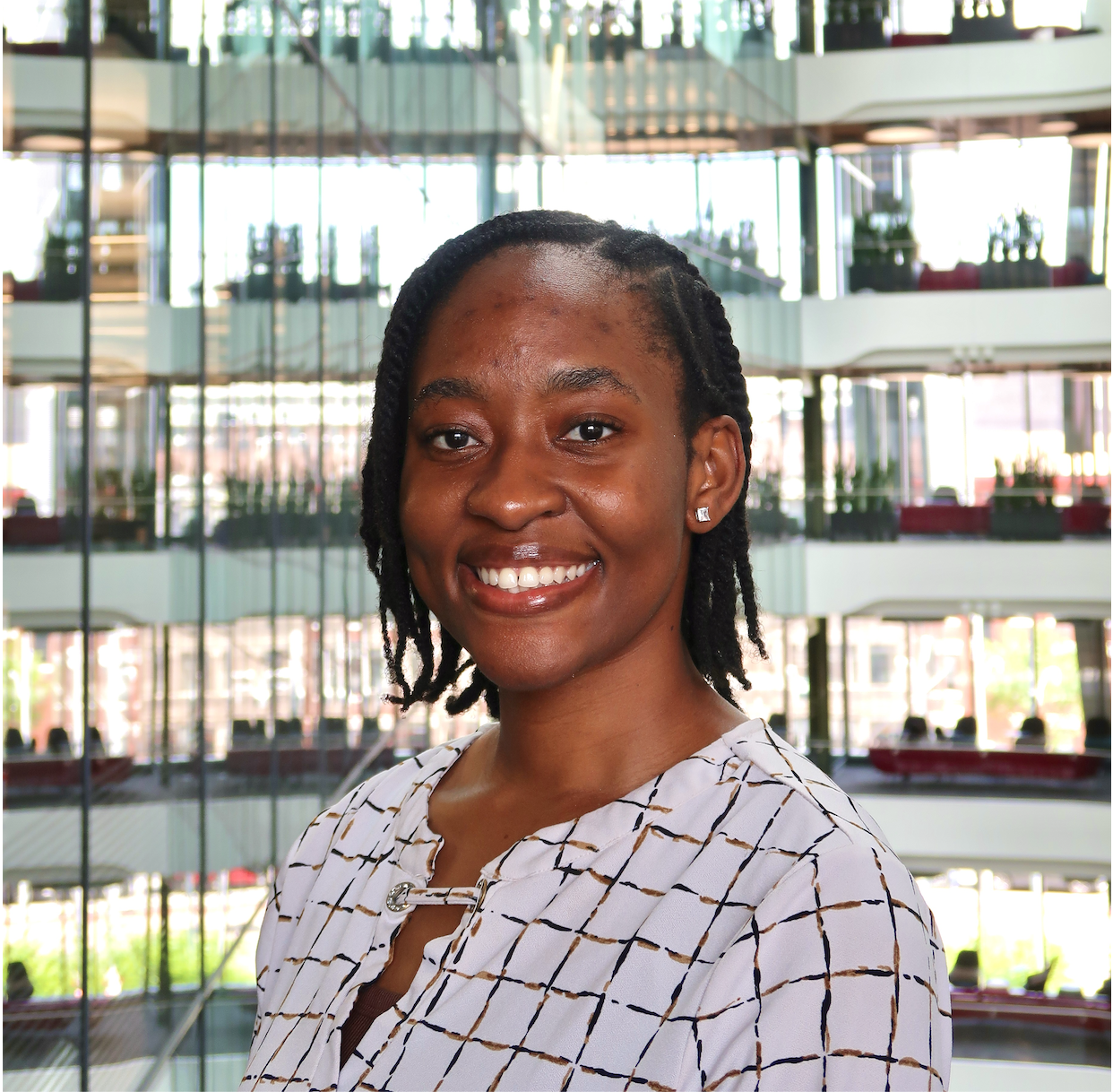A smiling African American woman with short twists hair, wearing a white blouse with a checkered pattern, standing in front of a modern, multi-story office building with glass windows.