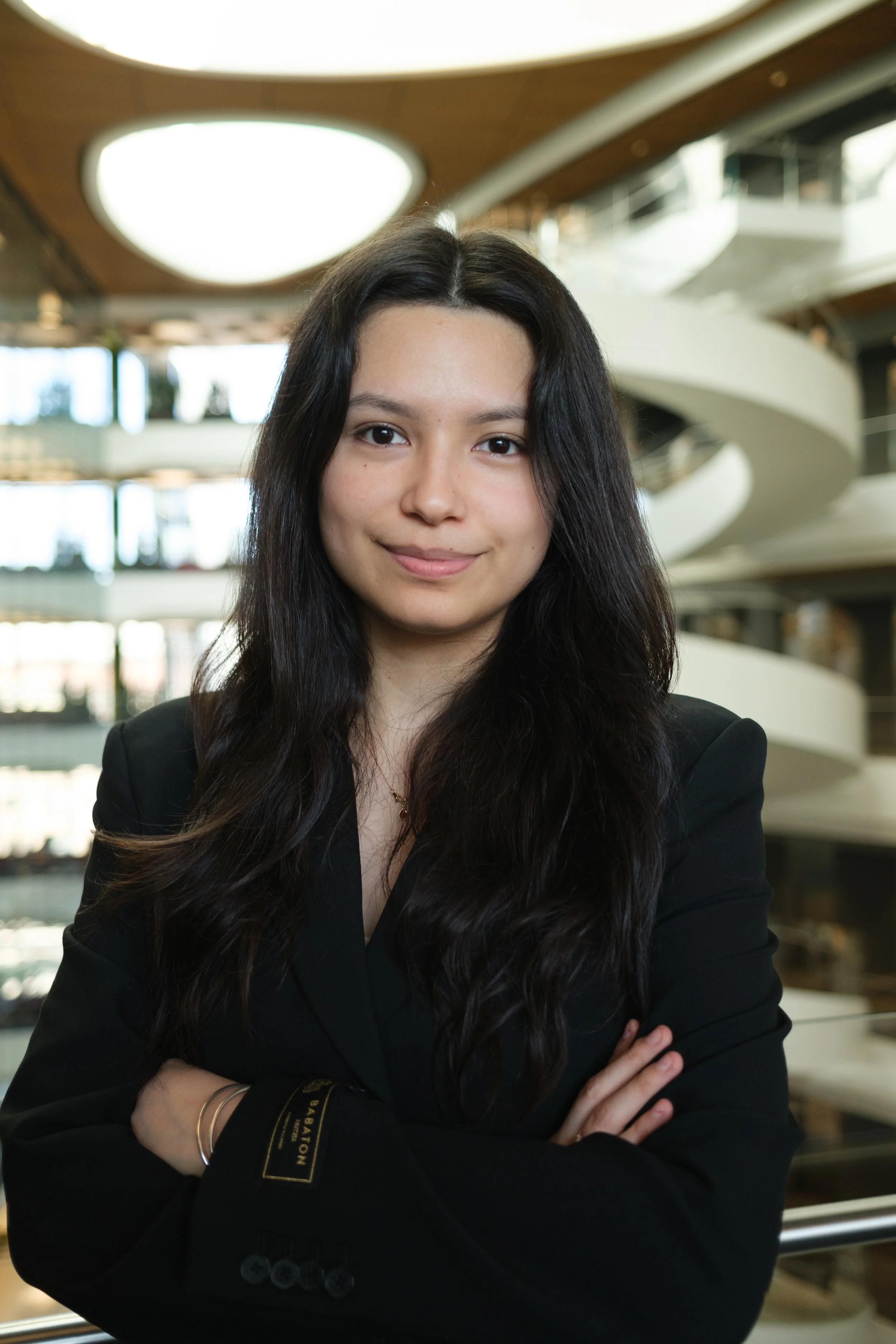 A young woman with long dark hair standing inside a modern building with a spiral staircase in the background, wearing a black blazer and crossing her arms.