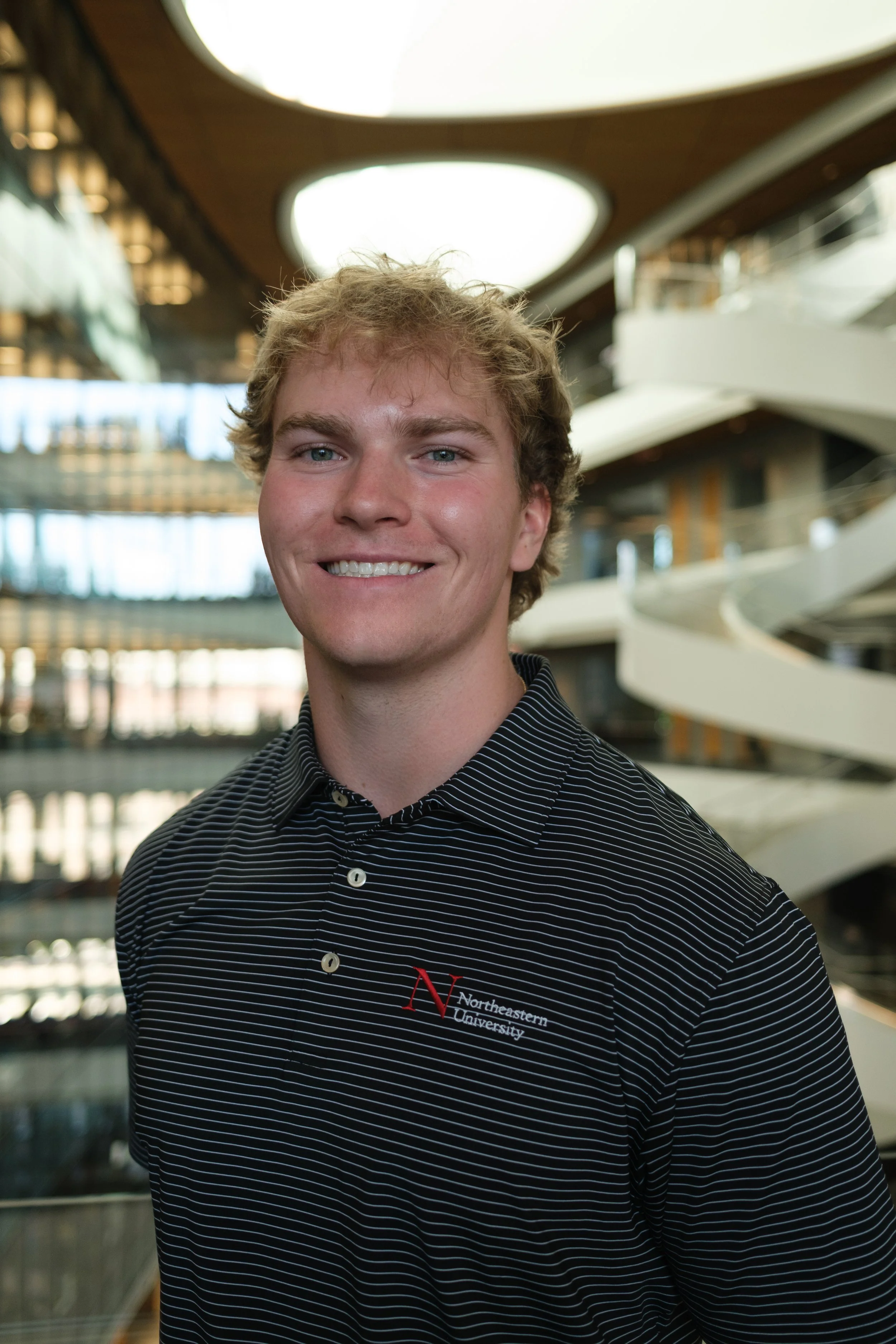 Smiling young man wearing a Northeastern University polo shirt, standing in front of a modern building with interior balconies and a spiral staircase.