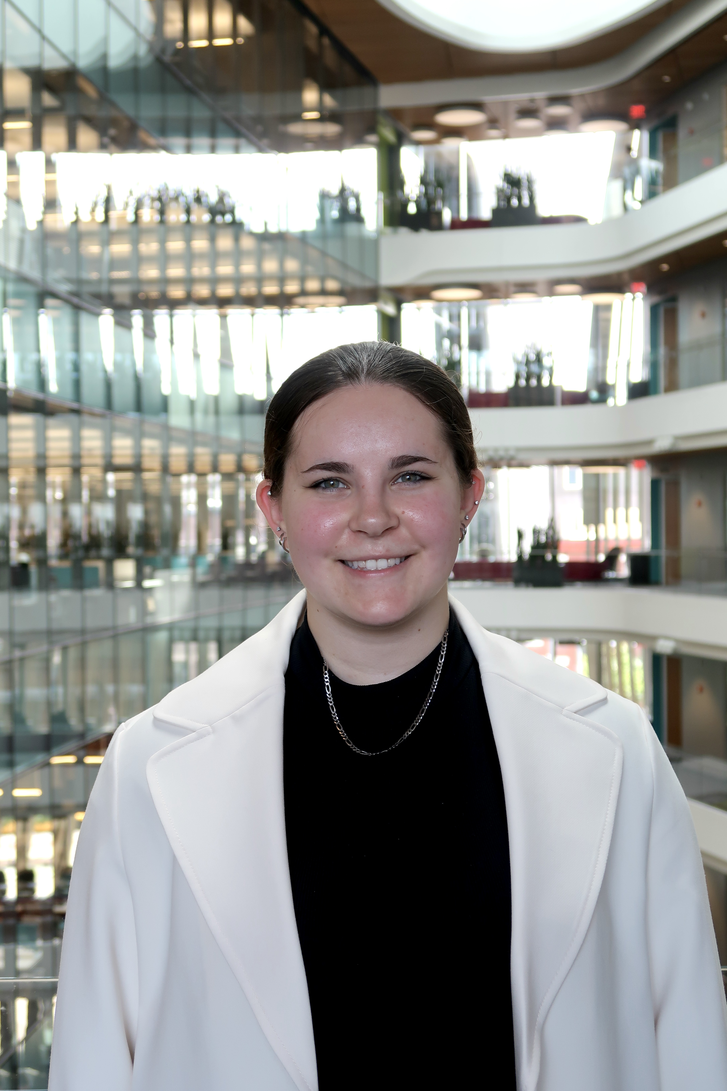 A smiling woman with dark hair pulled back, wearing a white blazer and black top with a silver chain necklace, standing inside a modern multi-story building with glass walls and open balconies.