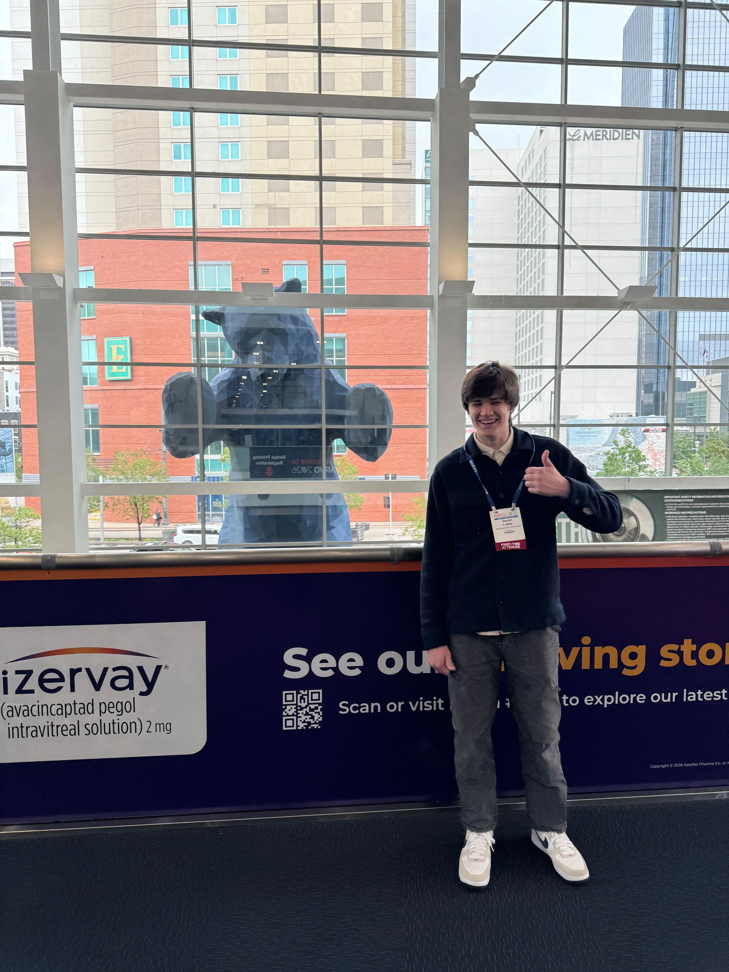 A young man standing in front of a large window with a cityscape, smiling and giving a thumbs-up gesture, wearing a black jacket, gray pants, and white sneakers.