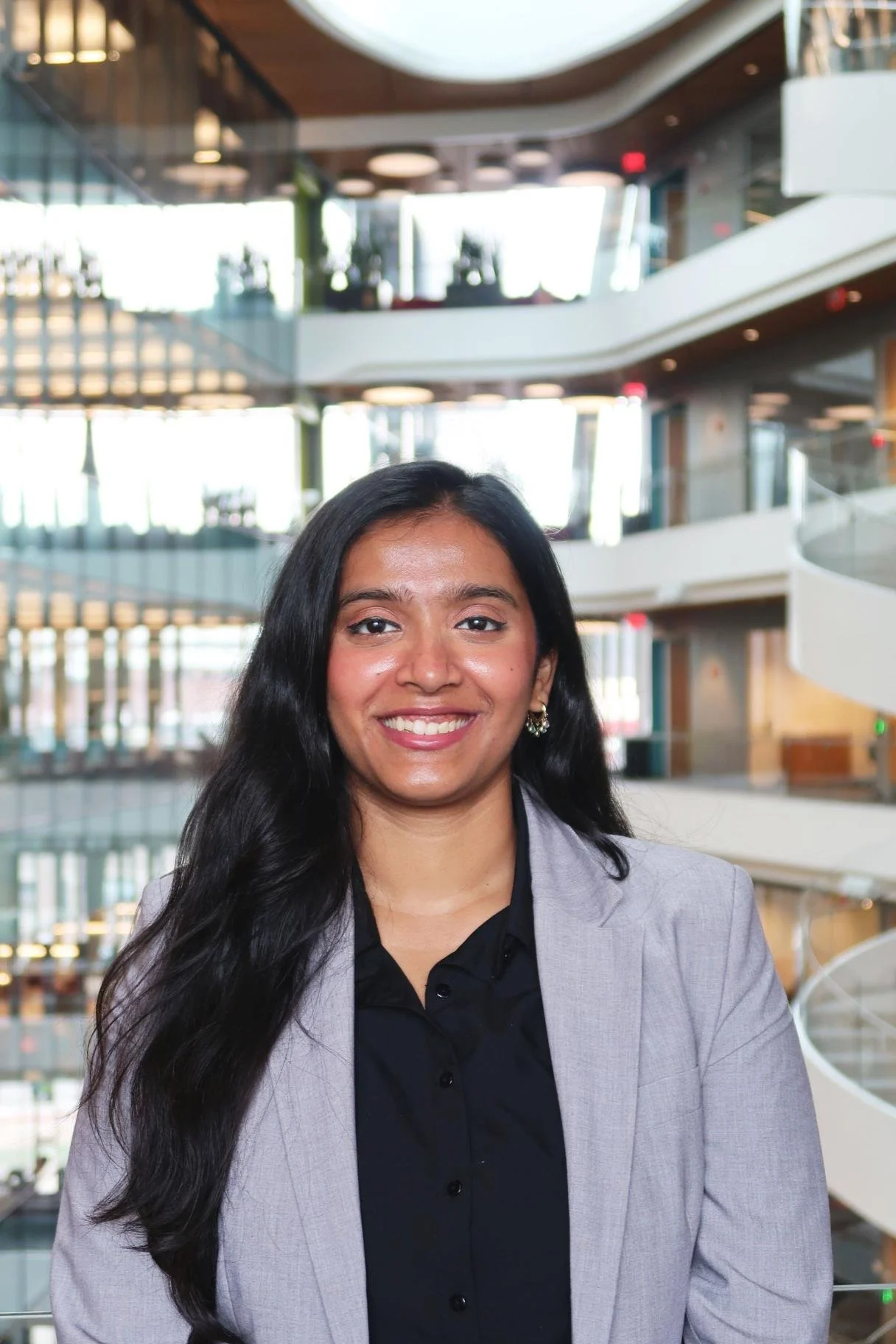 Portrait of a smiling woman with long dark hair, wearing a blazer and earrings, in a modern multi-story building with glass walls and staircases.