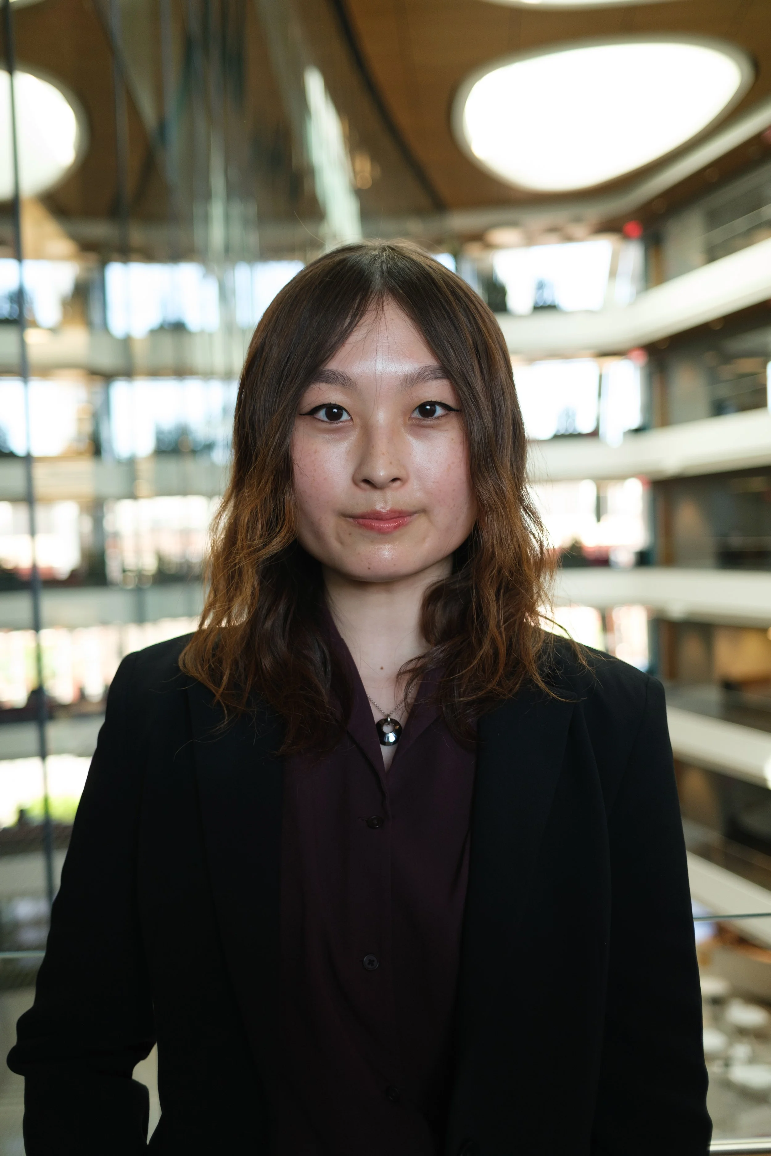 A young woman with wavy brown hair, wearing a black blazer and maroon blouse, stands inside a modern building with glass walls and multiple floors in the background.