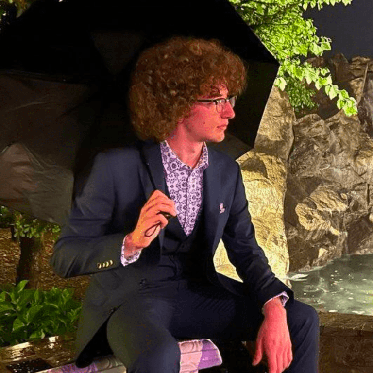 A man with curly hair, glasses, and a suit sitting outdoors next to rocks and plants, holding an umbrella.