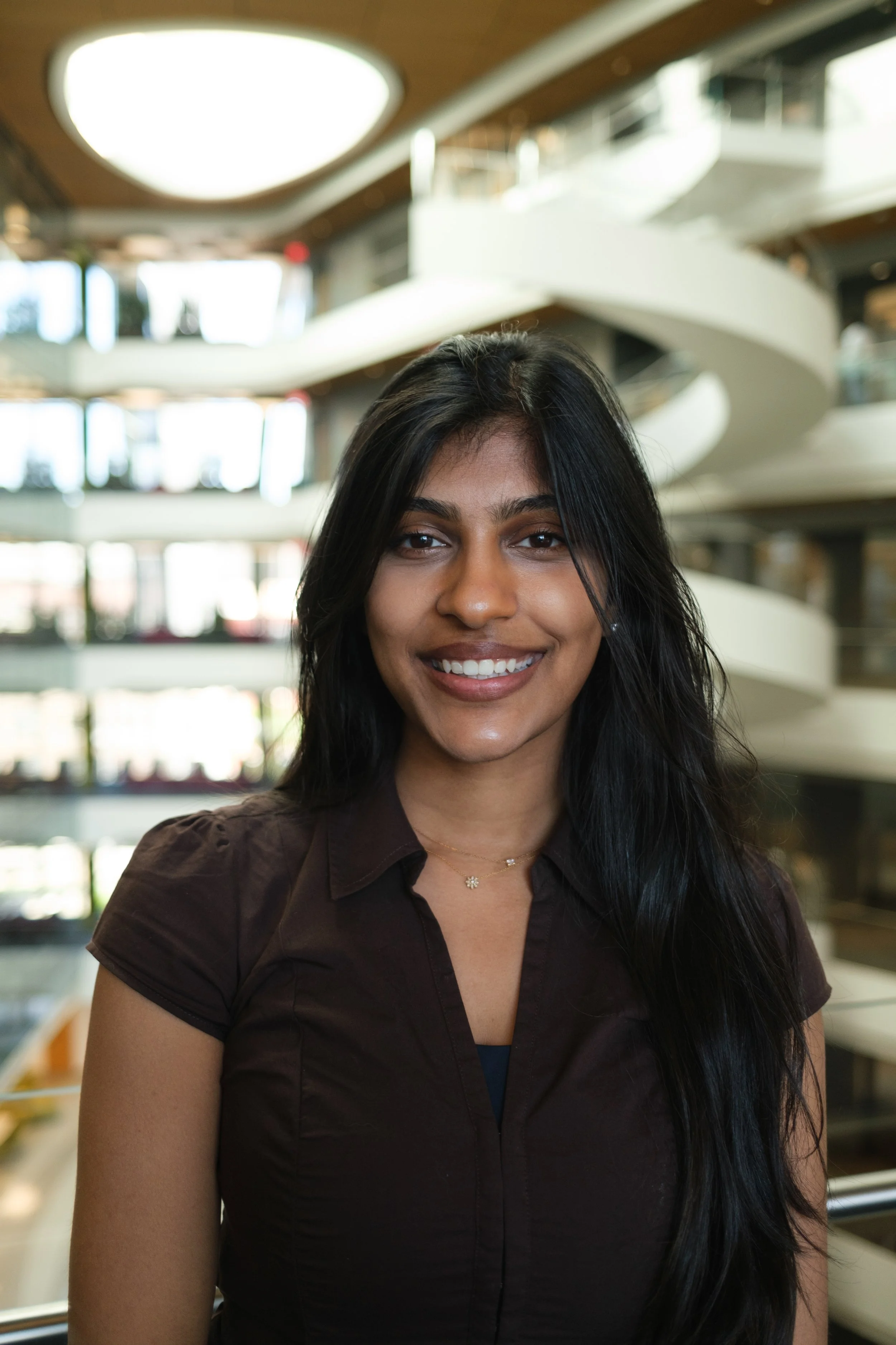 A young woman with long black hair and a brown shirt smiling indoors at a modern building with curved white balconies.