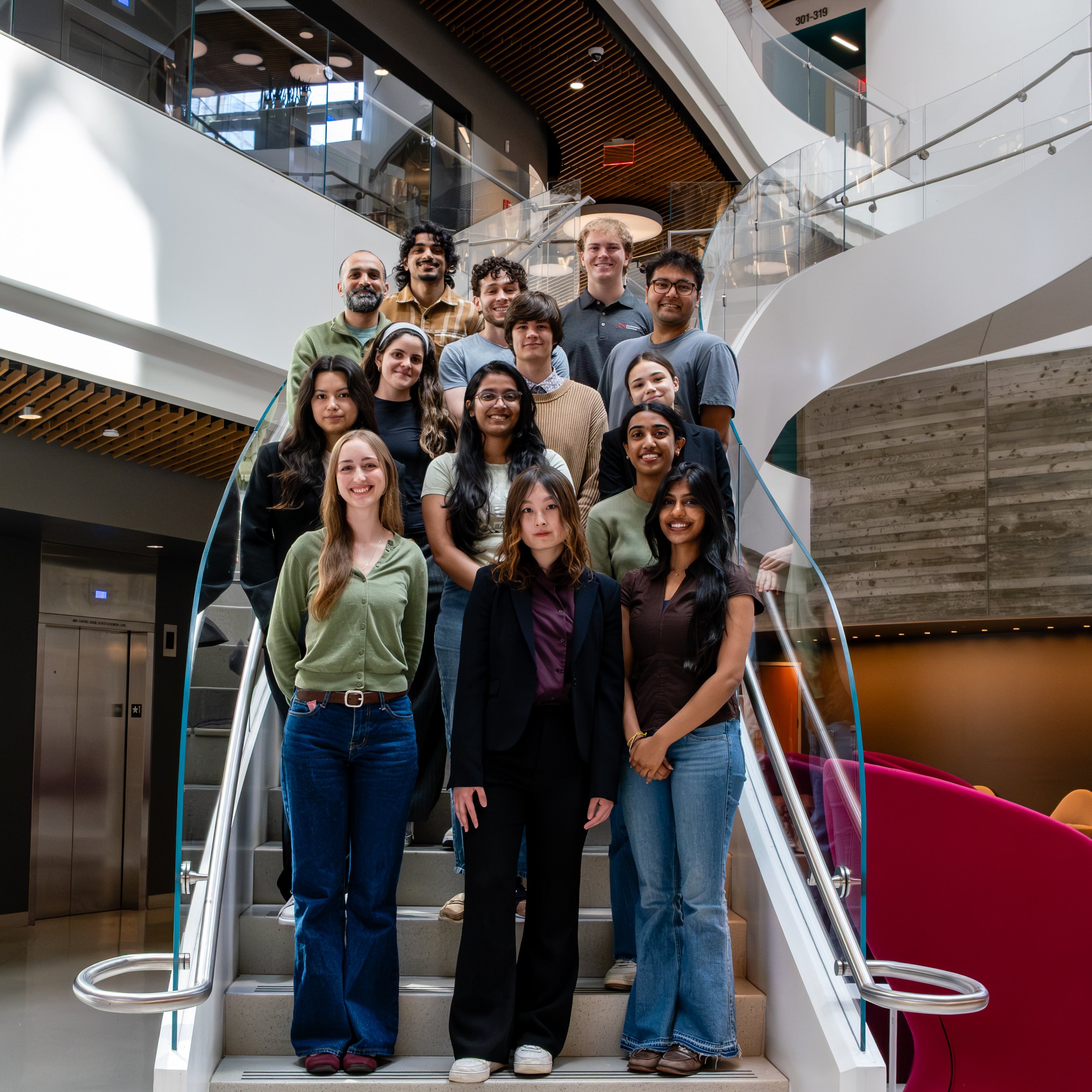 Group of diverse young adults standing on staircase inside modern building, smiling at camera.