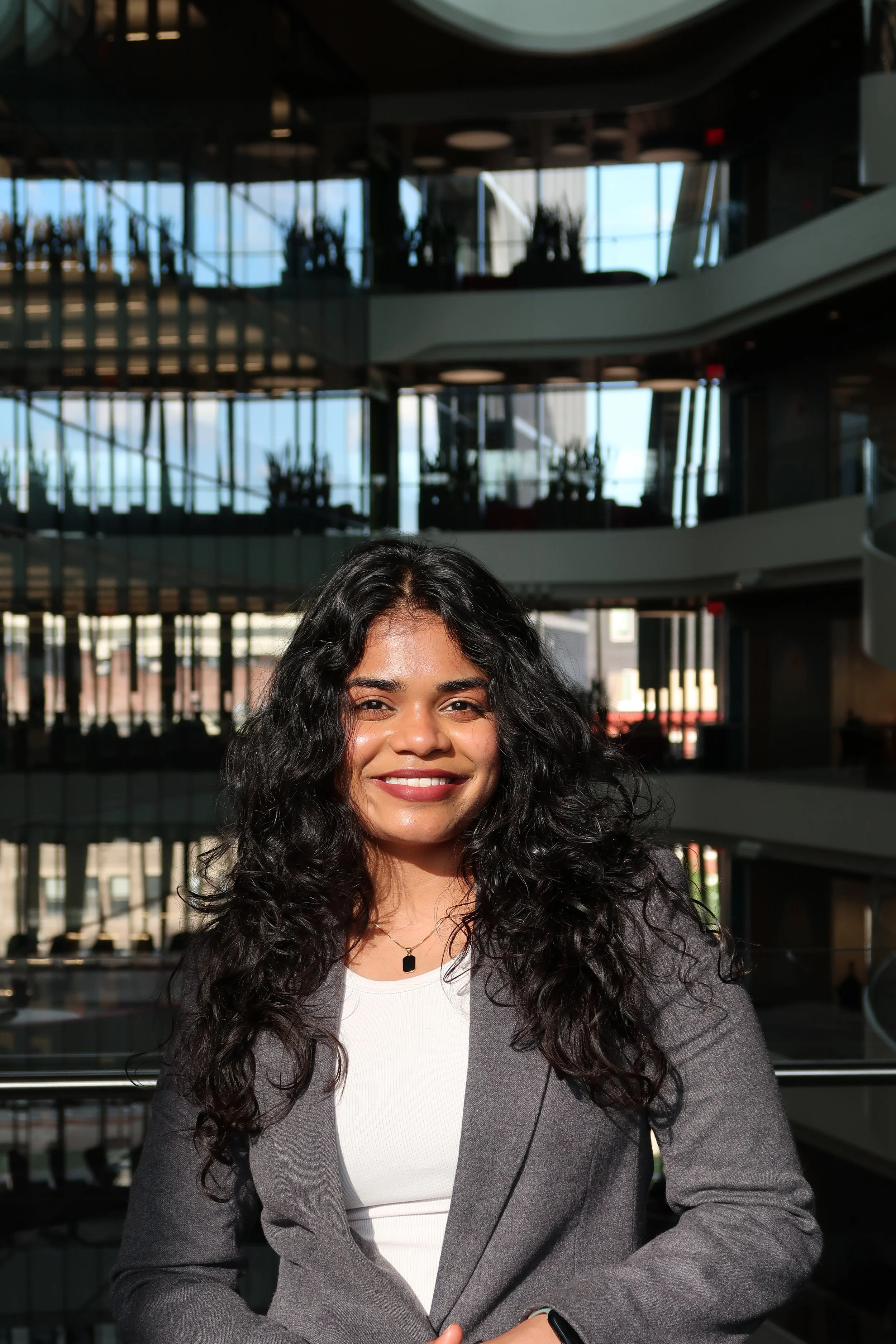 A woman with curly dark hair smiling, wearing a white top and gray blazer, standing in front of a modern building with glass windows and multiple floors.