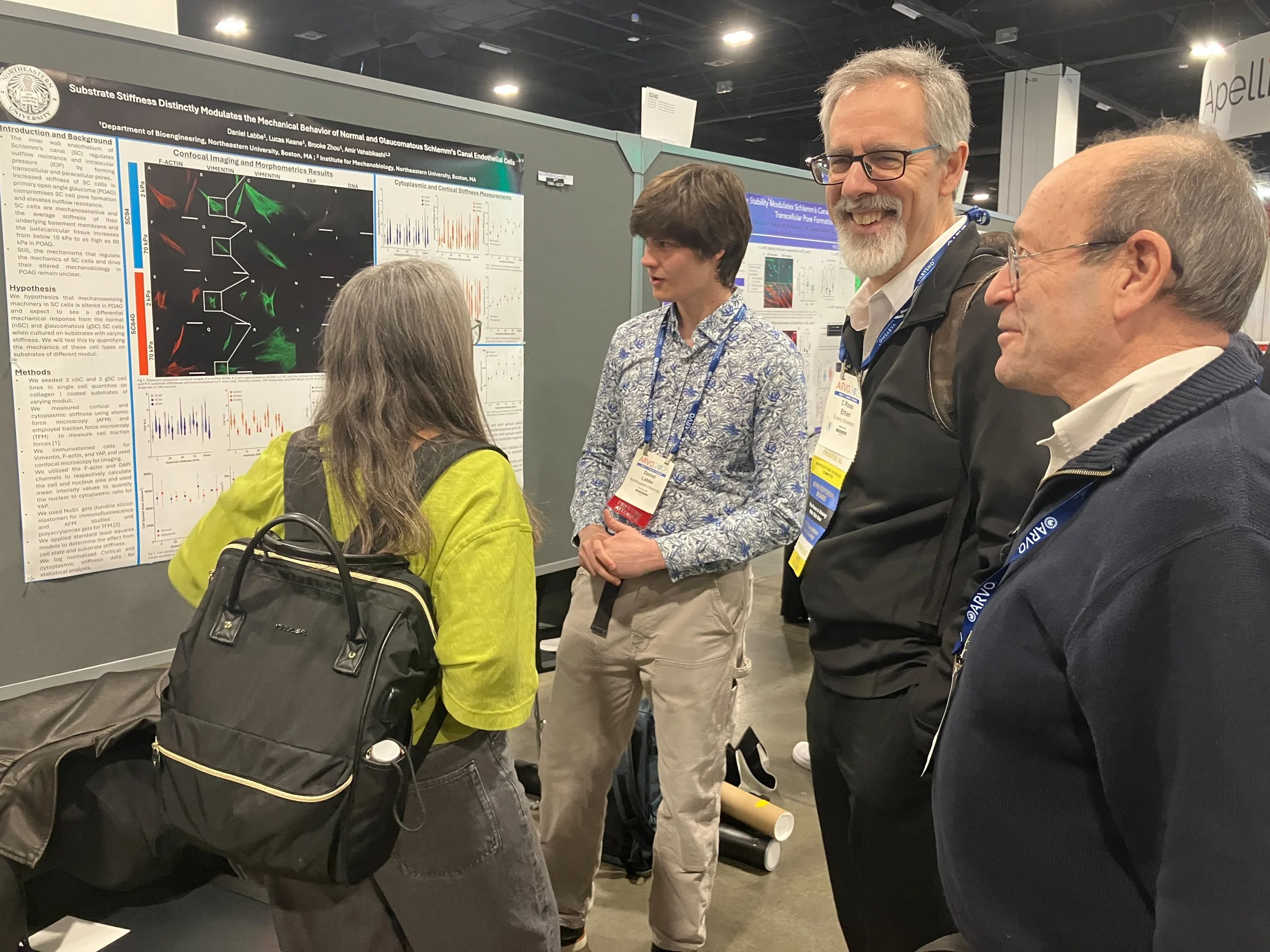 A group of three people, two men and one woman, standing at a conference poster presentation. They are engaged in a discussion about the scientific poster, which contains charts, graphs, and text. The woman, wearing a yellow top and gray pants, is speaking to the two men. The men are listening attentively, one with glasses, a gray beard, and a black jacket, the other with glasses and a patterned shirt. The conference setting has gray walls and other poster displays in the background.