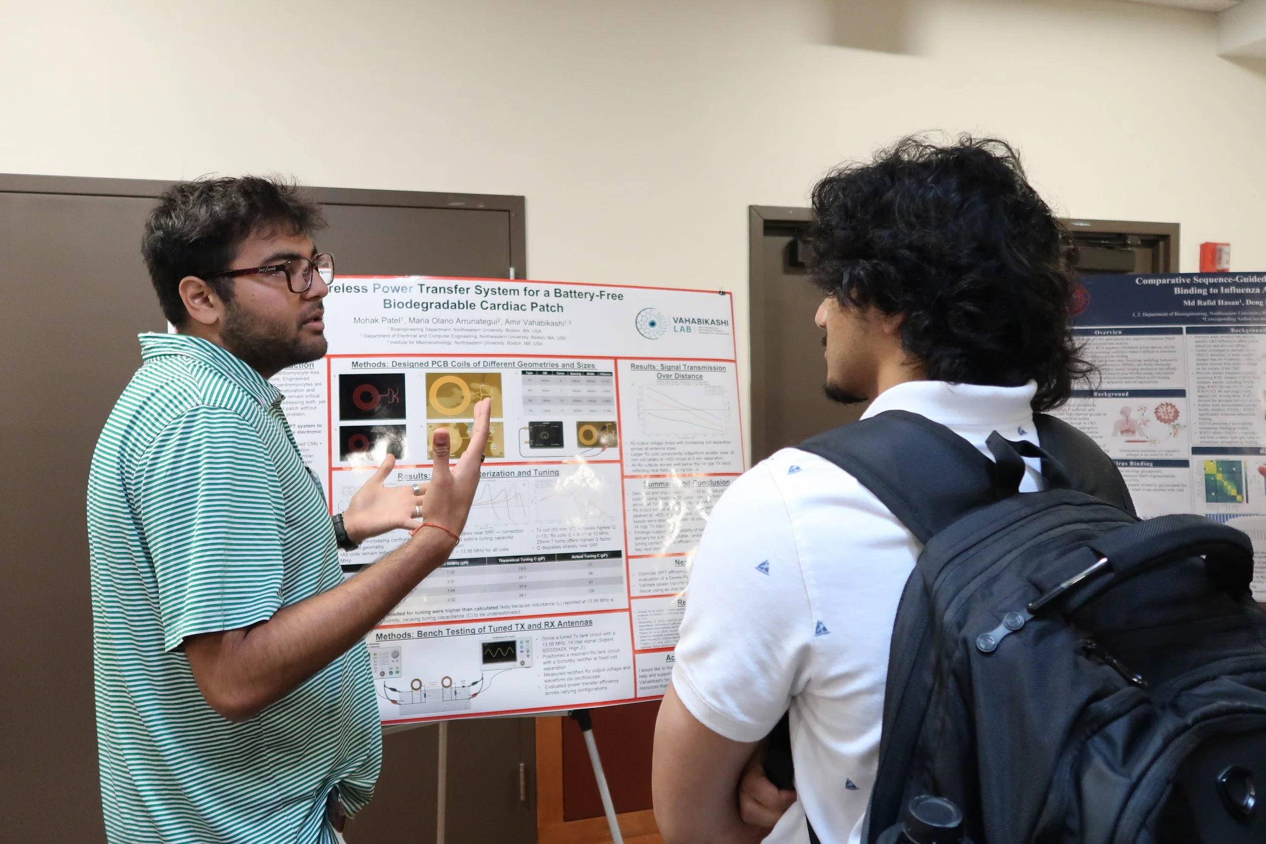 Two young men are standing in front of a research poster, engaged in discussion. One man is gesturing with his hands, while the other is listening attentively. The poster contains text, images, and graphs related to scientific research.