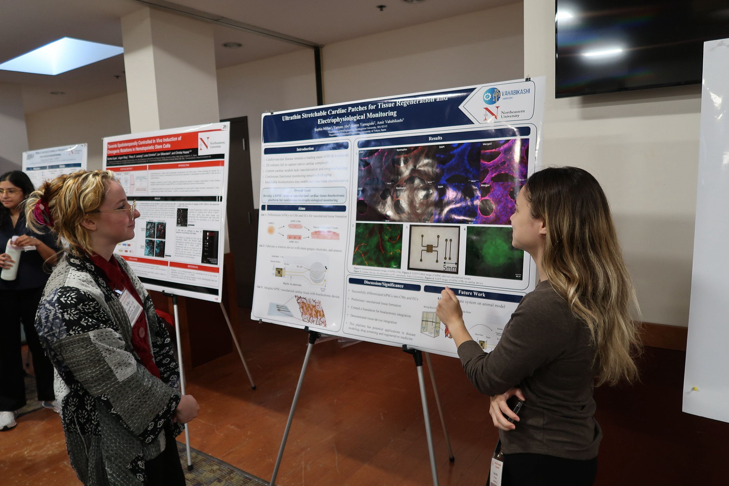 Two women engage in conversation at a scientific conference. One woman is looking at a research poster titled 'Ultrathin Stretchable Cardiac Patches for Tissue Regeneration and Electrophysiological Monitoring' by researchers from North Eastern University and VAHABIKASHI Laboratory. The poster displays images, diagrams, and data related to cardiac tissue engineering.