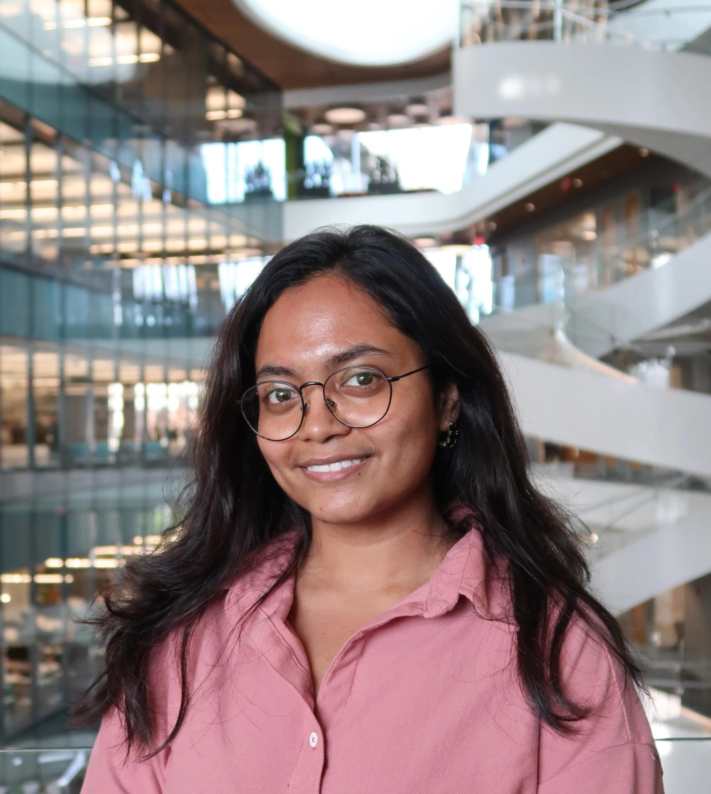 Close-up of a woman wearing glasses and a pink button-up shirt, smiling, inside a modern, multi-story building with glass and metal architecture.