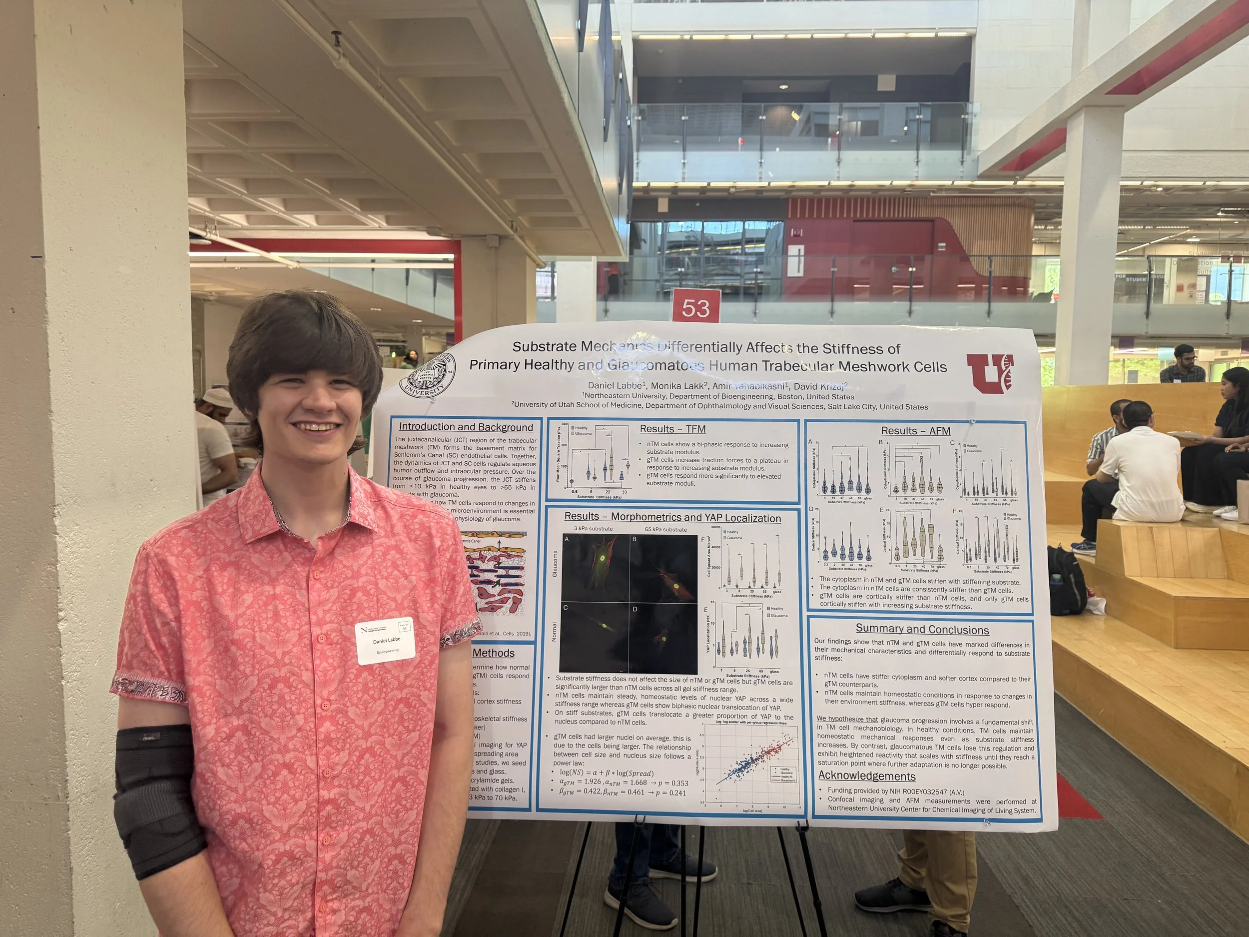 A young man wearing a pink shirt and black wrist brace standing in front of a science research poster at an academic conference, smiling at the camera.