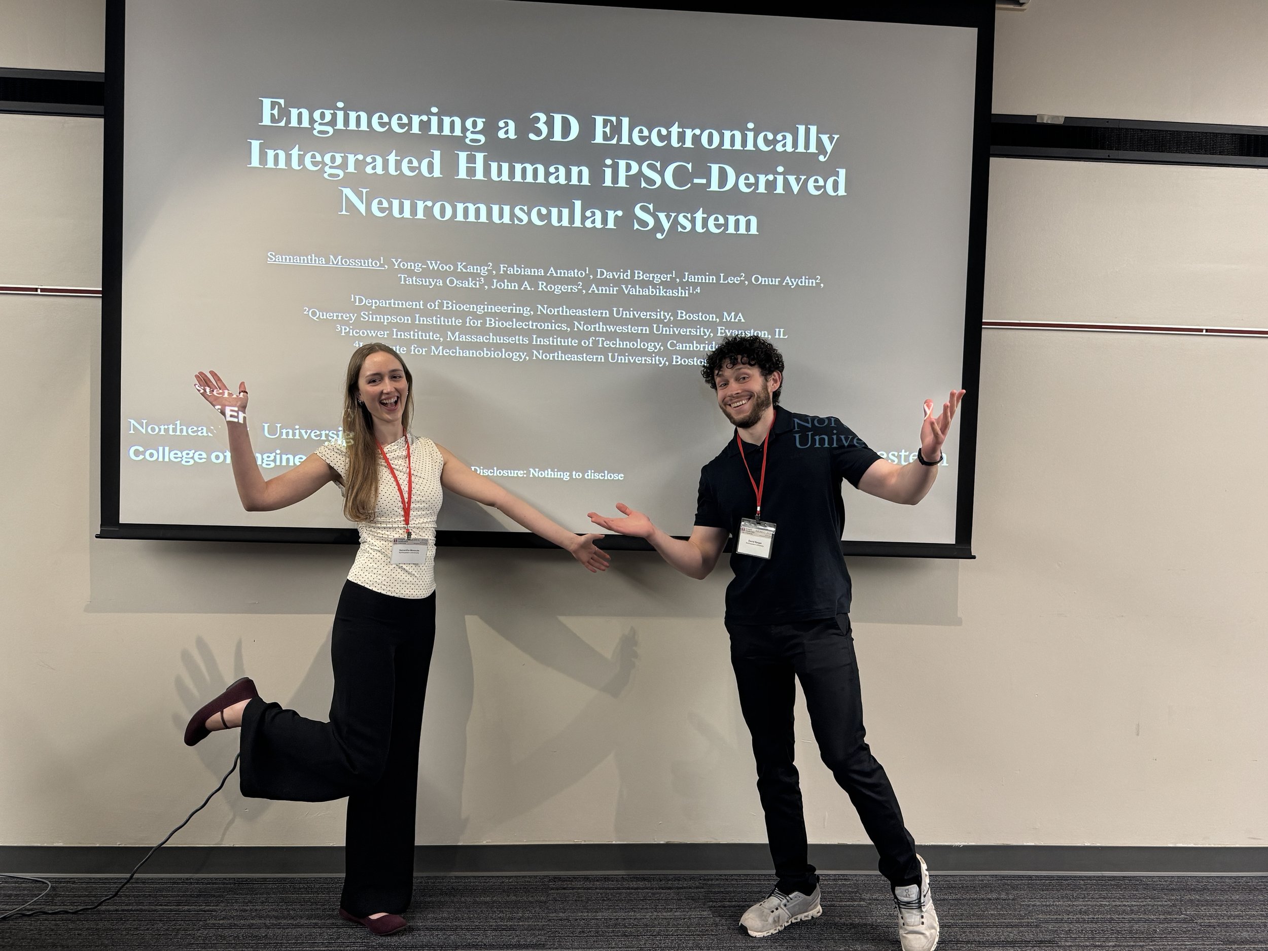 Two people posing playfully in front of a presentation screen displaying a scientific research title. The woman on the left is standing with one foot raised and smiling, while the man on the right is standing with his arms open, also smiling. The presentation slide behind them shows the title 'Engineering a 3D Electronically Integrated Human iPSC-Derived Neuromuscular System' and lists the authors and their affiliations.