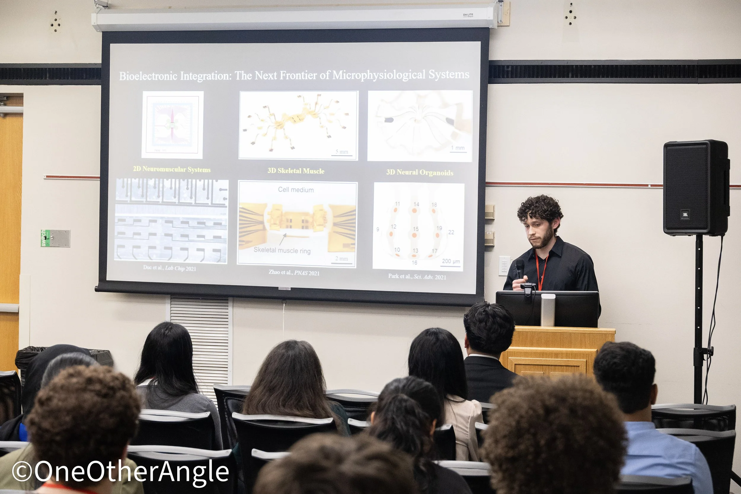 A classroom or conference room with an audience watching a presenter giving a lecture on bioelectronic integration and microphysiological systems. The slide on the screen displays images and titles such as '2D Neuromuscular Systems,' '3D Skeletal Muscle,' and '3D Neural Organoids.' The presenter is a young man with curly hair wearing a black shirt, standing next to a large speaker.