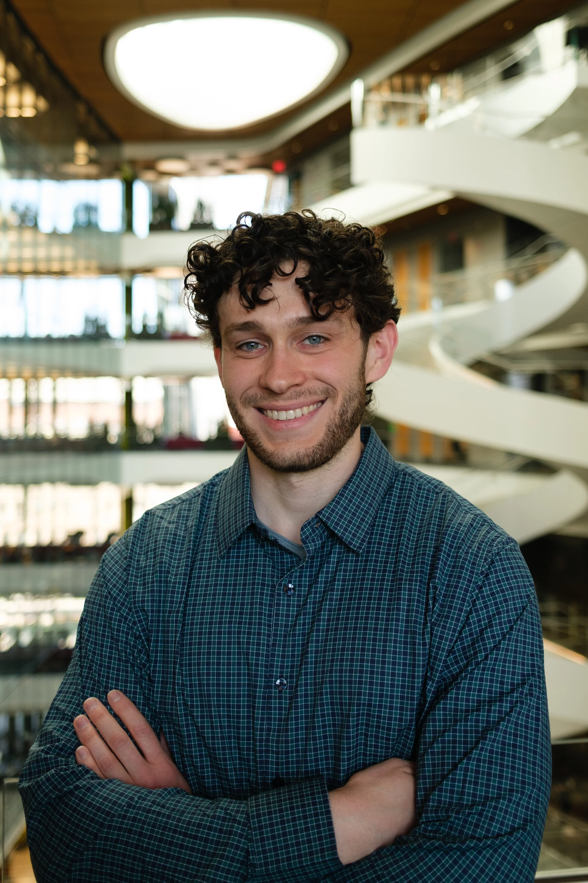 Headshot of a young man with curly dark hair, blue eyes, and a beard, smiling while standing with arms crossed in a modern, multi-story building with curved staircases and large windows.