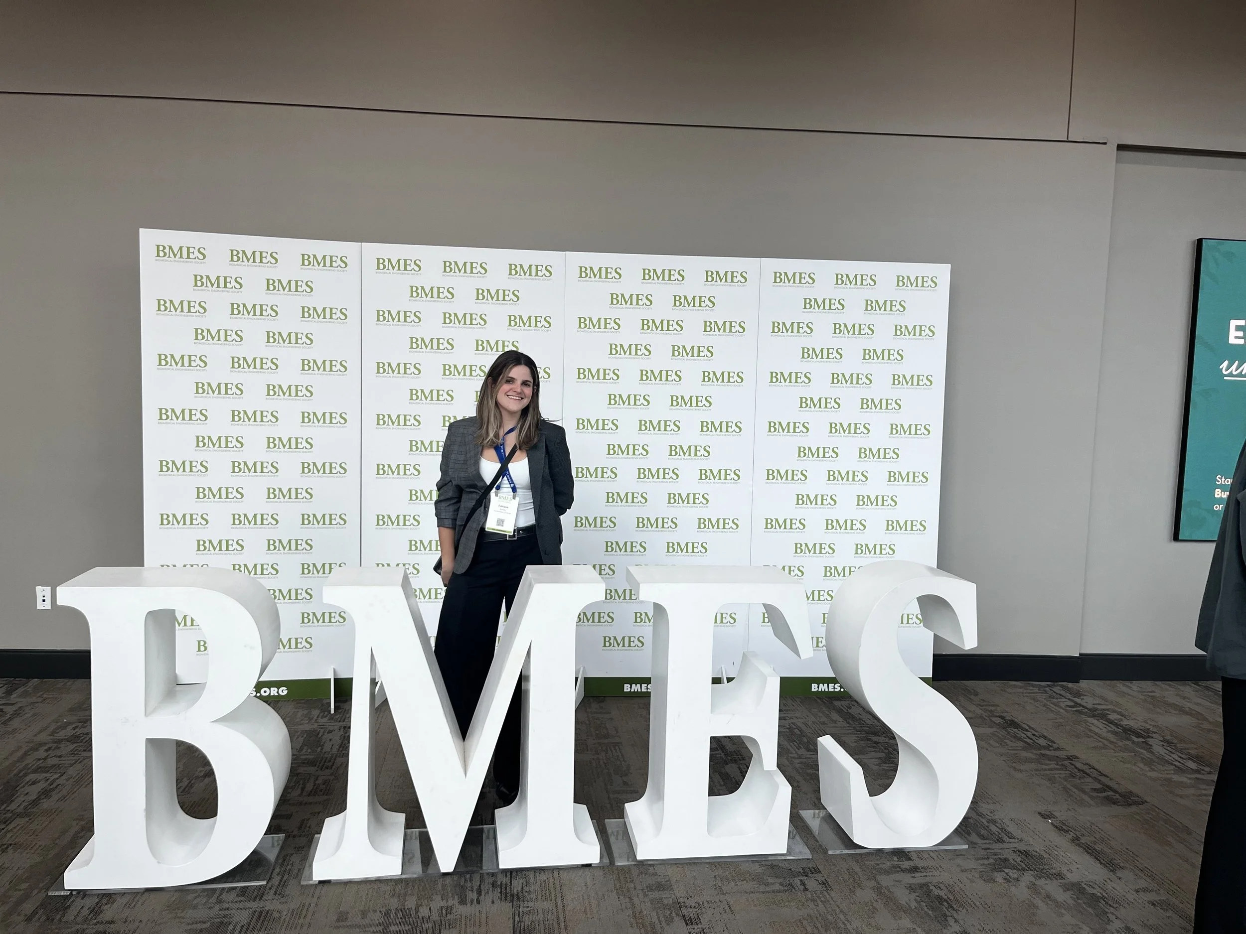 Young woman smiling, wearing a blazer, standing in front of a backdrop with the repeated logo 'BMES' at a professional conference or event. Large white letters spelling 'BVES' are in front of her on the floor.