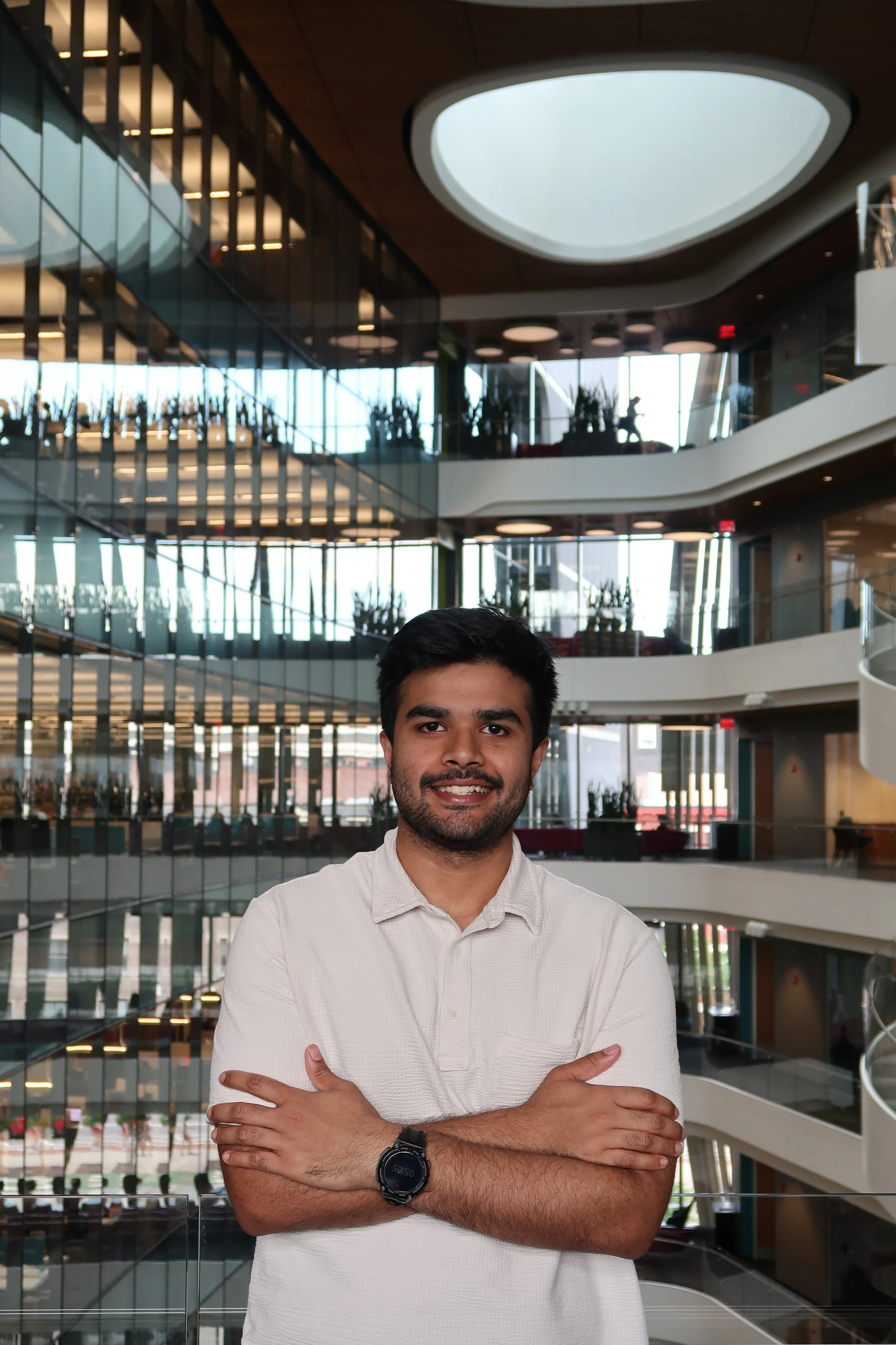 A man with dark hair and a beard wearing a white shirt and a black watch, standing inside a modern office building with curved balconies, glass walls, and potted plants.