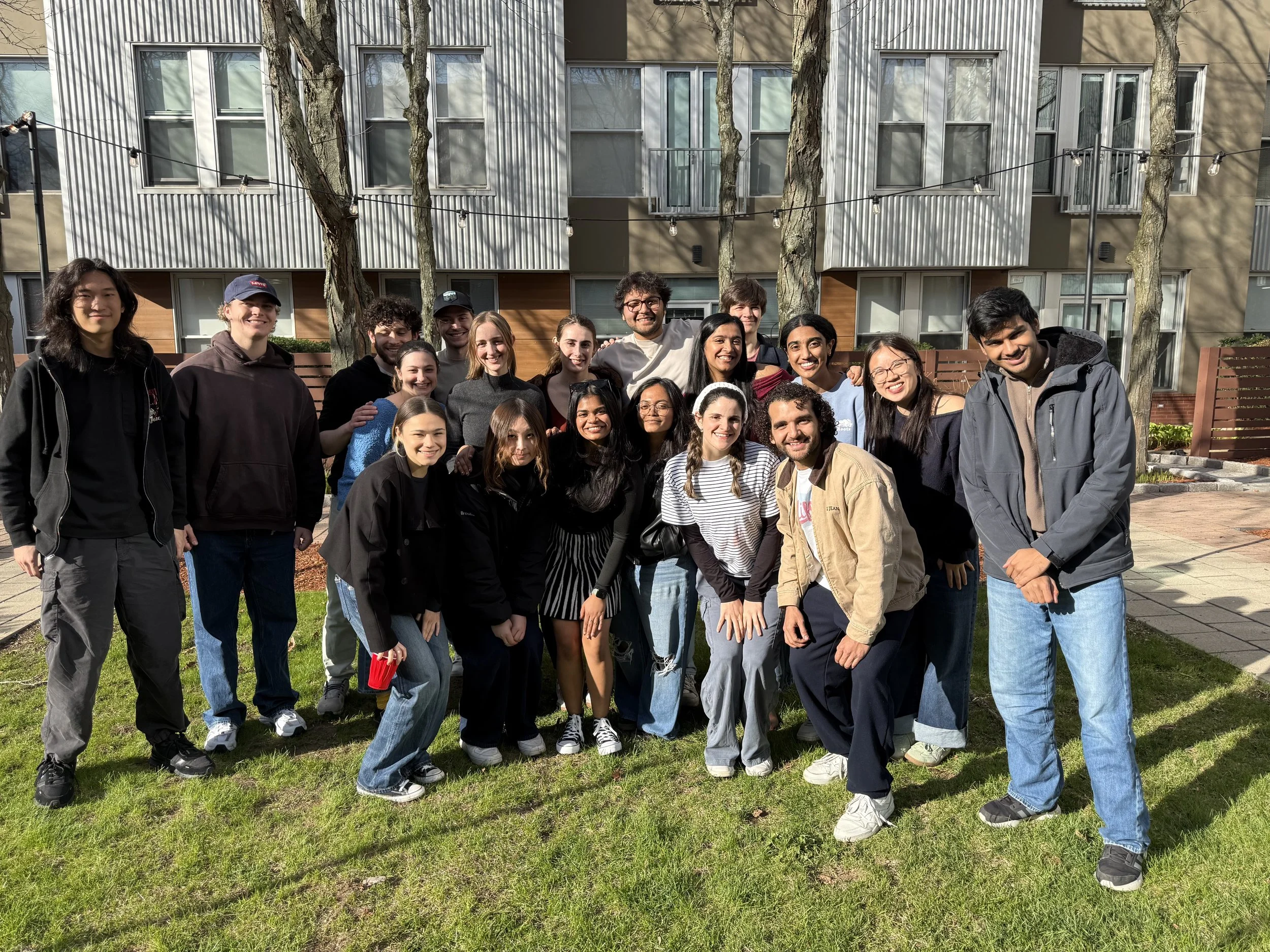 A group of diverse young people smiling and posing together outdoors in front of a modern apartment building with trees and string lights.