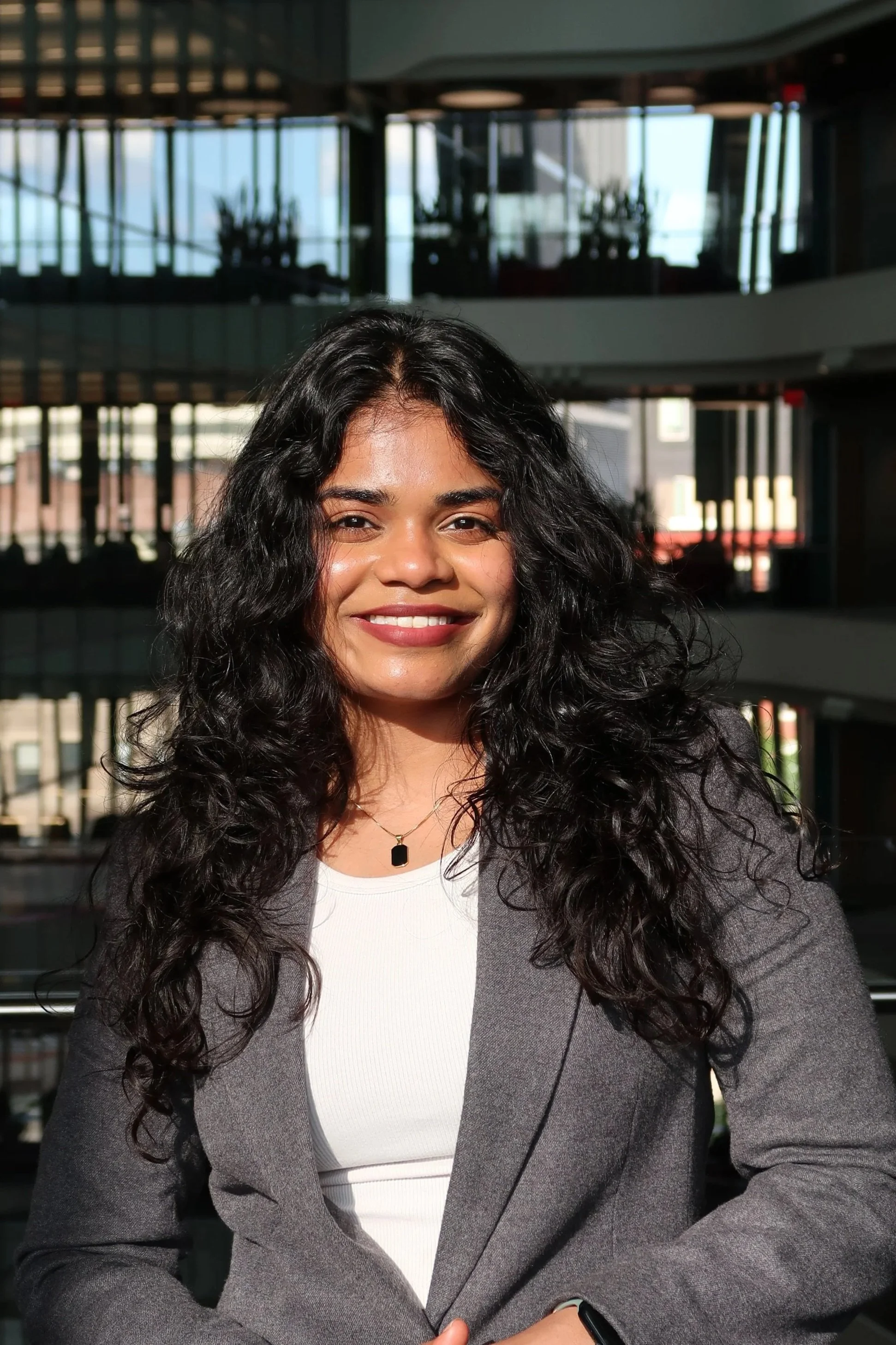 A woman with long curly black hair smiling, wearing a white top, gray blazer, gold necklace with a square pendant, and a smartwatch, in a modern office building with glass walls and plants in the background.