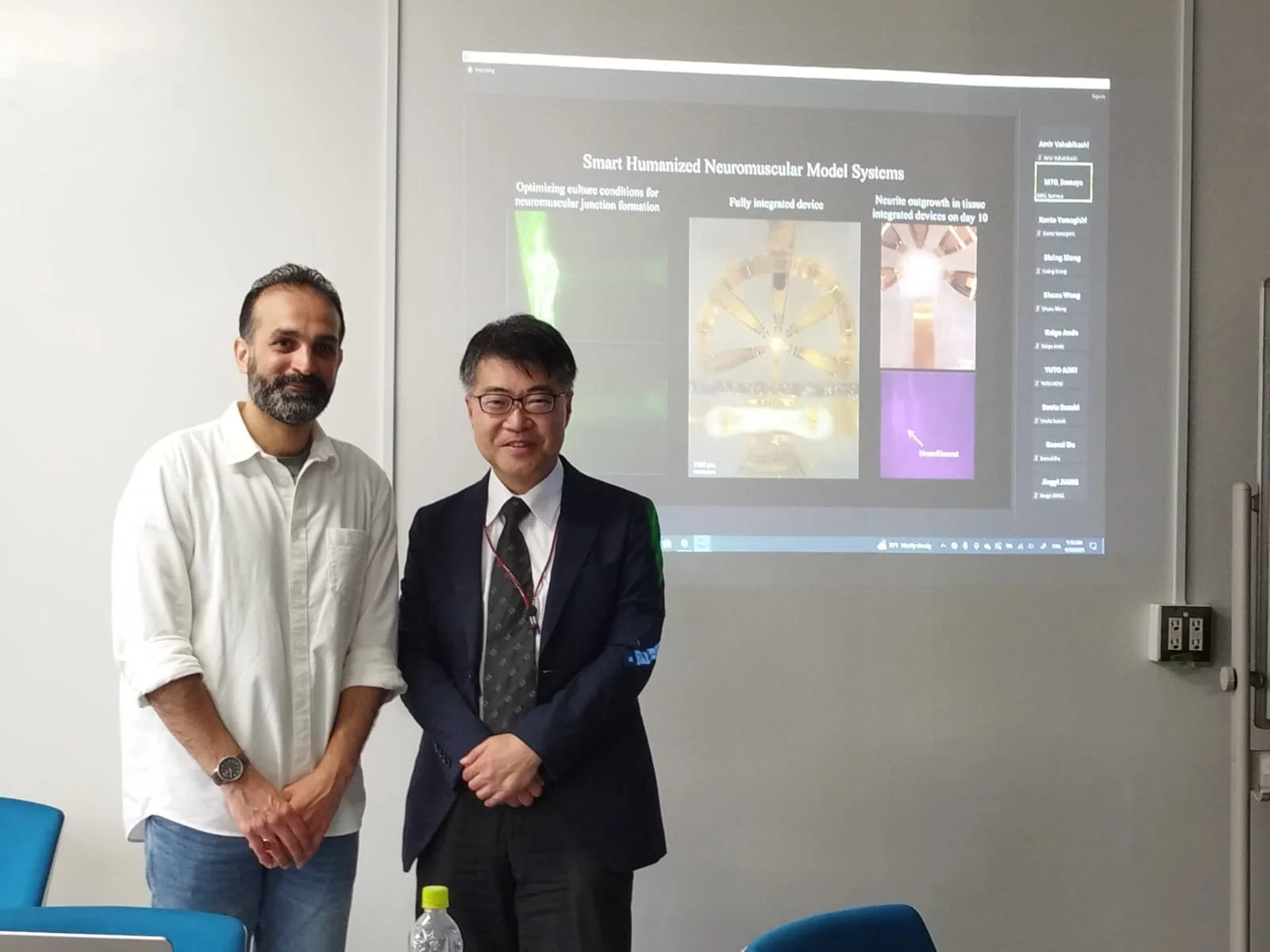 Two men standing in front of a presentation slide titled "Smart Humanized Neuromuscular Model Systems." One man is dressed casually in a white shirt and jeans, and the other is in a dark suit with glasses. A water bottle is on the table in front of them.