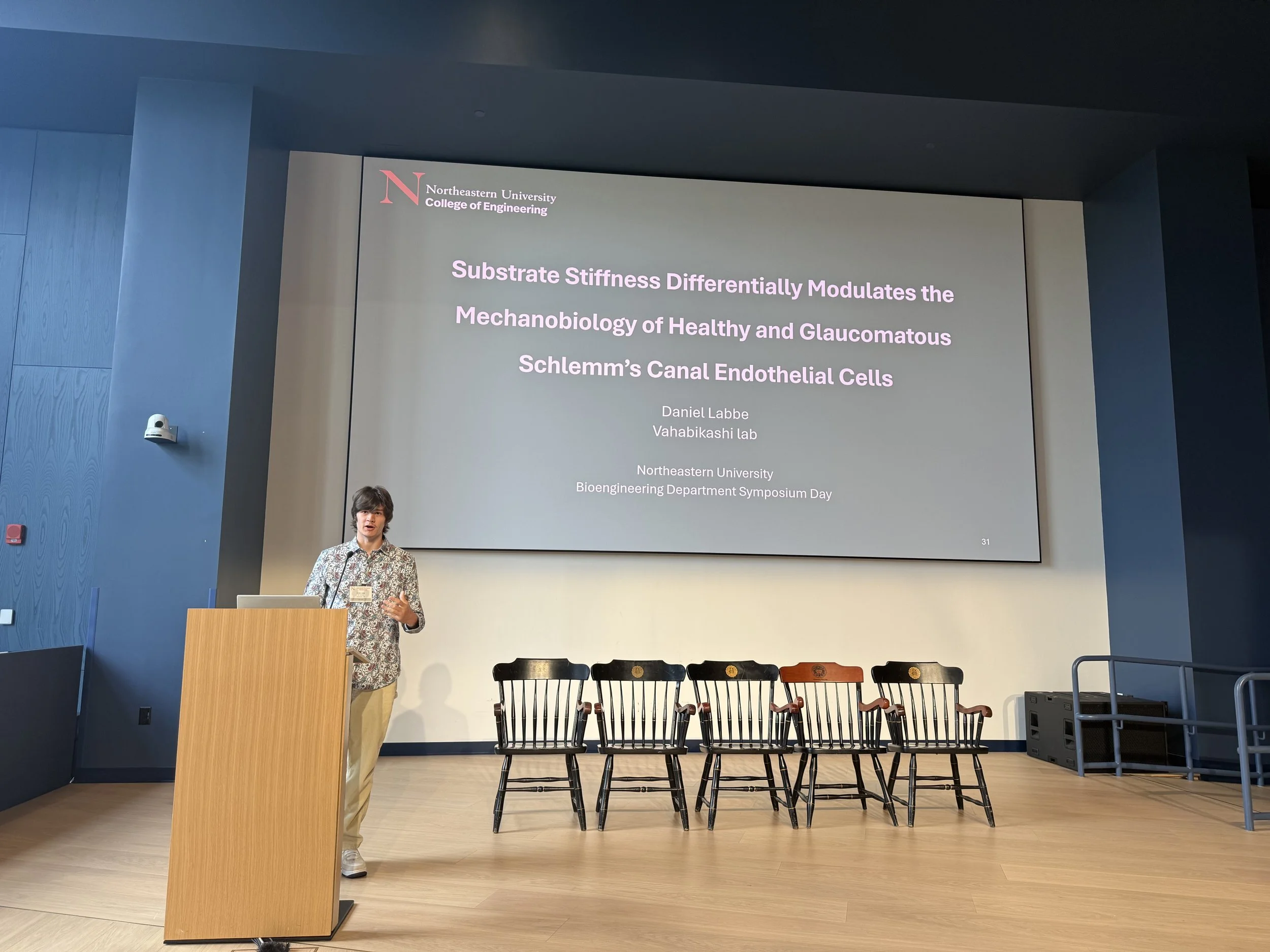 A person standing at a wooden podium giving a presentation in a large auditorium or conference room. A large screen behind them displays a slide titled 'Substrate Stiffness Differentially Modulates the Mechanobiology of Healthy and Glaucomatous Schlemm's Canal Endothelial Cells' with additional details about the speaker and the event at Northeastern University.