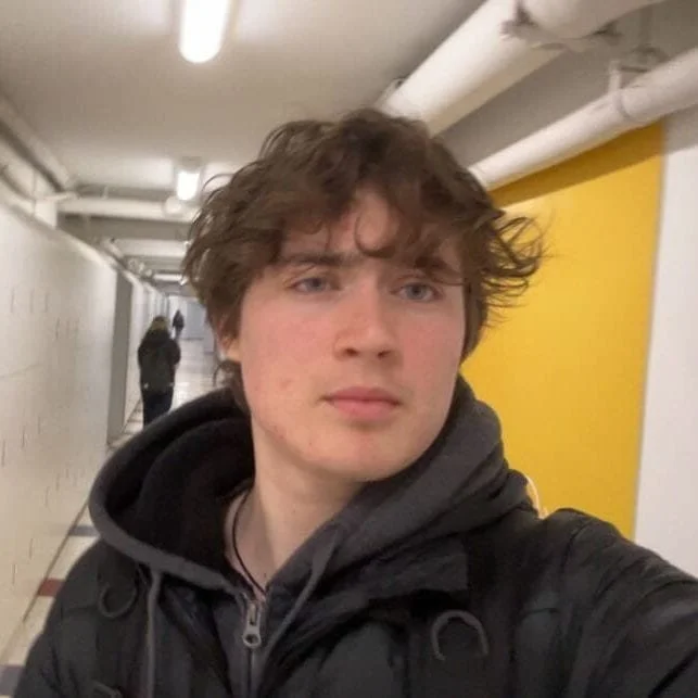 A young man with curly brown hair taking a selfie in a hallway with yellow and white walls.