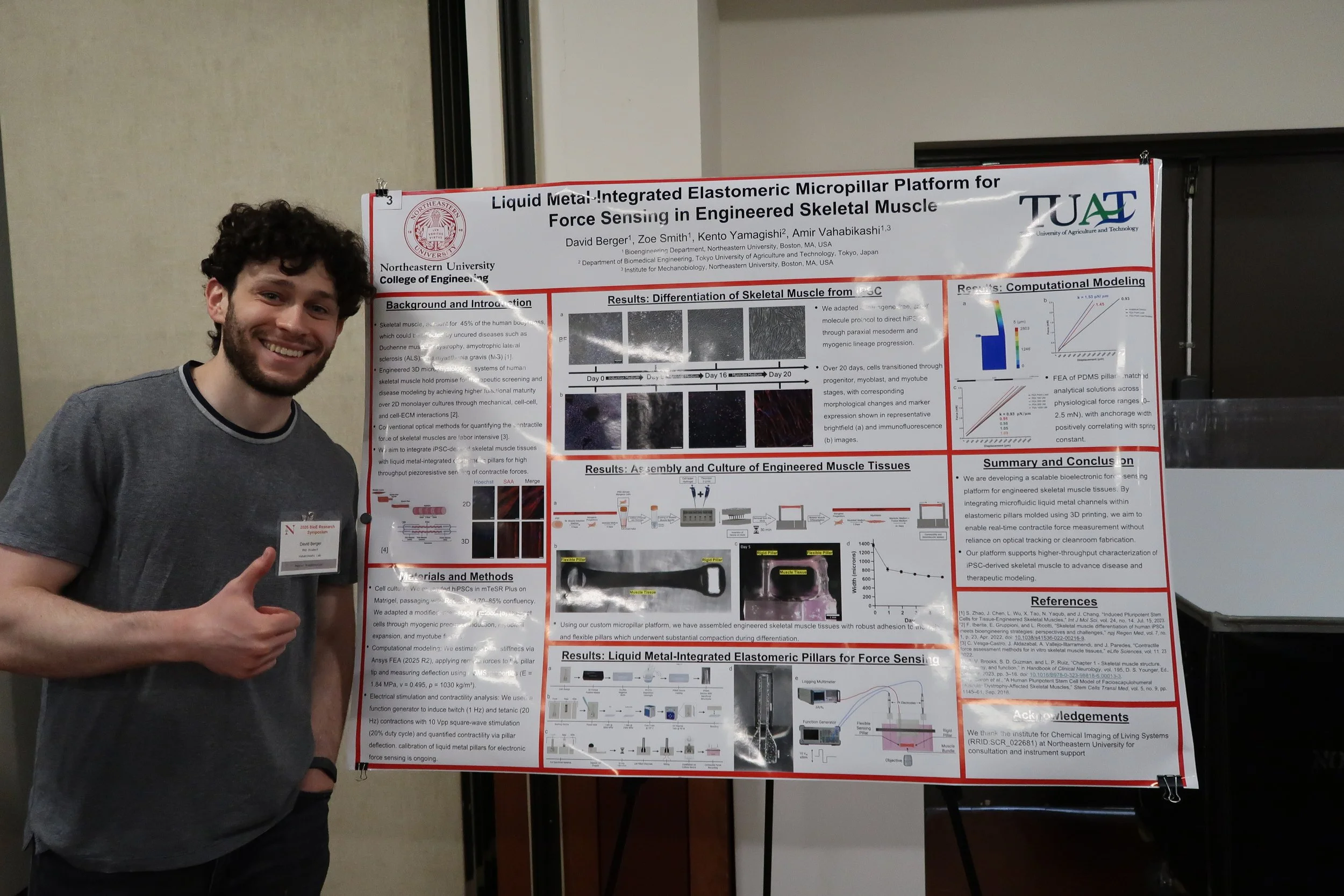 A young man with curly hair and a beard smiling and giving a thumbs up, standing next to a science research poster at a conference or event.