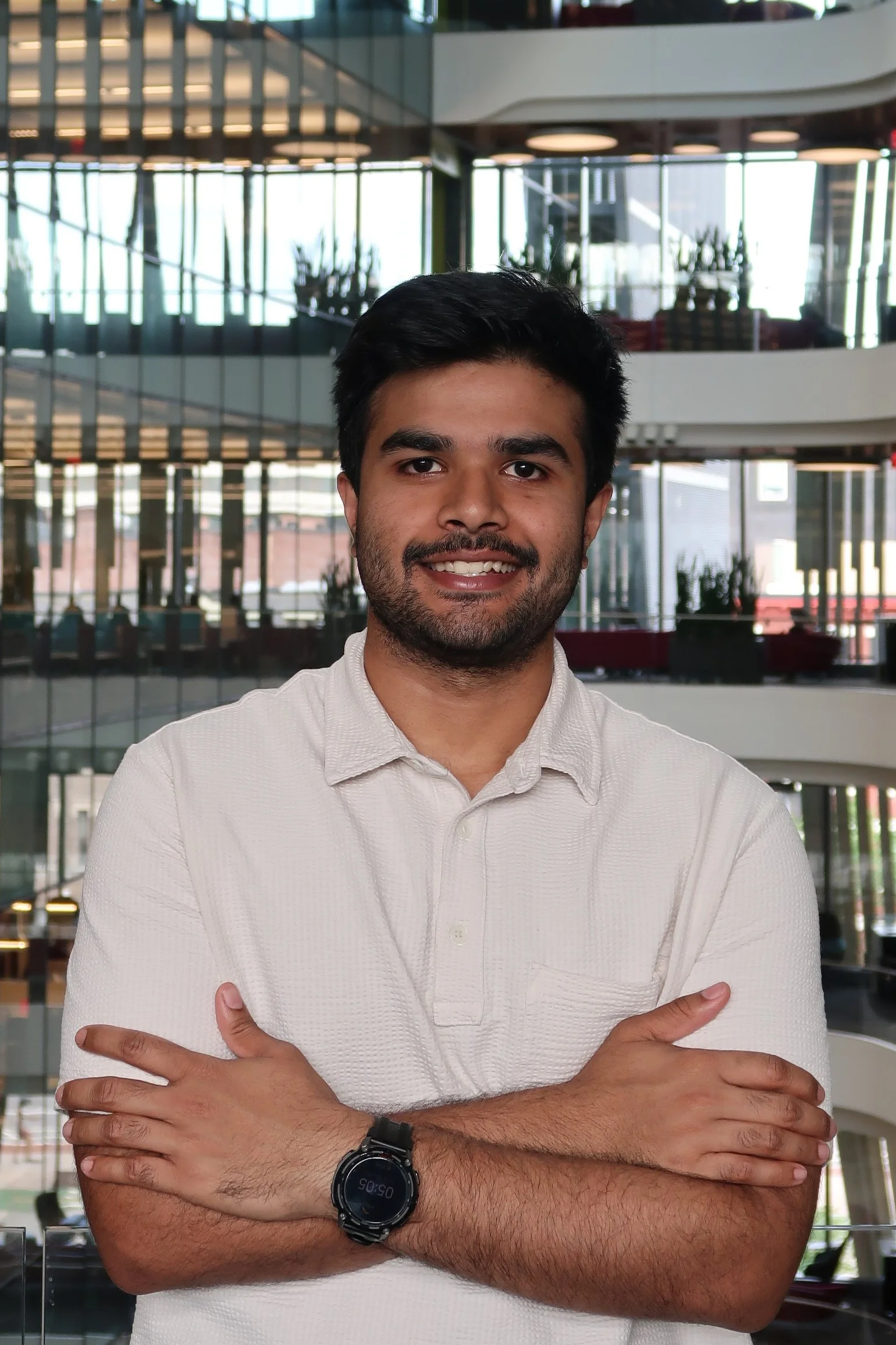 A smiling young man with dark hair and a beard, wearing a white shirt, standing in front of a large glass window with a modern, multi-story building interior background, arms crossed.