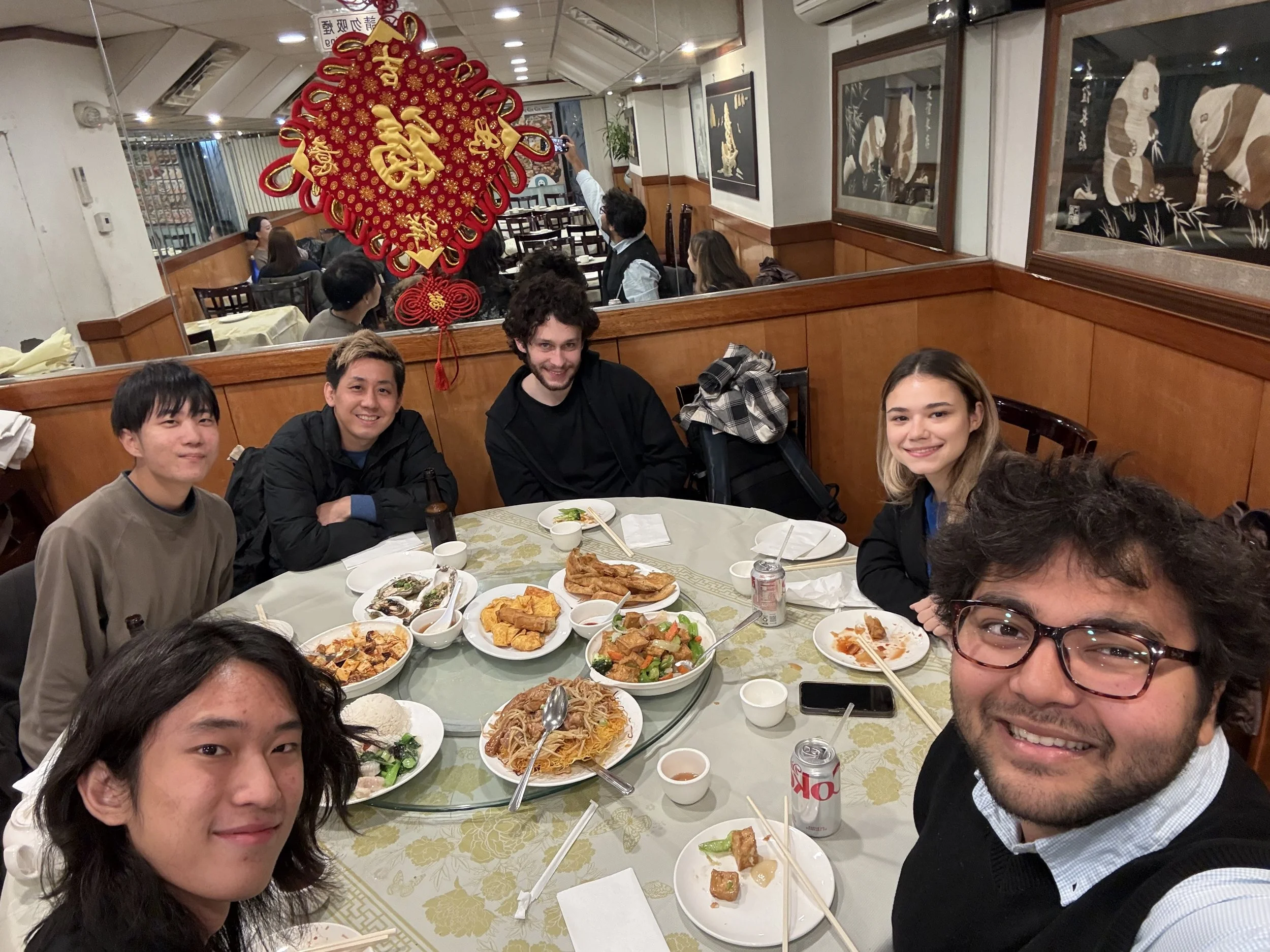 Group of people at a restaurant enjoying a meal with dishes like noodles, spring rolls, and vegetables on a round table. Some of them are smiling, and there are decorative red and gold Chinese New Year ornaments hanging from the ceiling. The restaurant has wooden paneling and framed artwork on the walls.