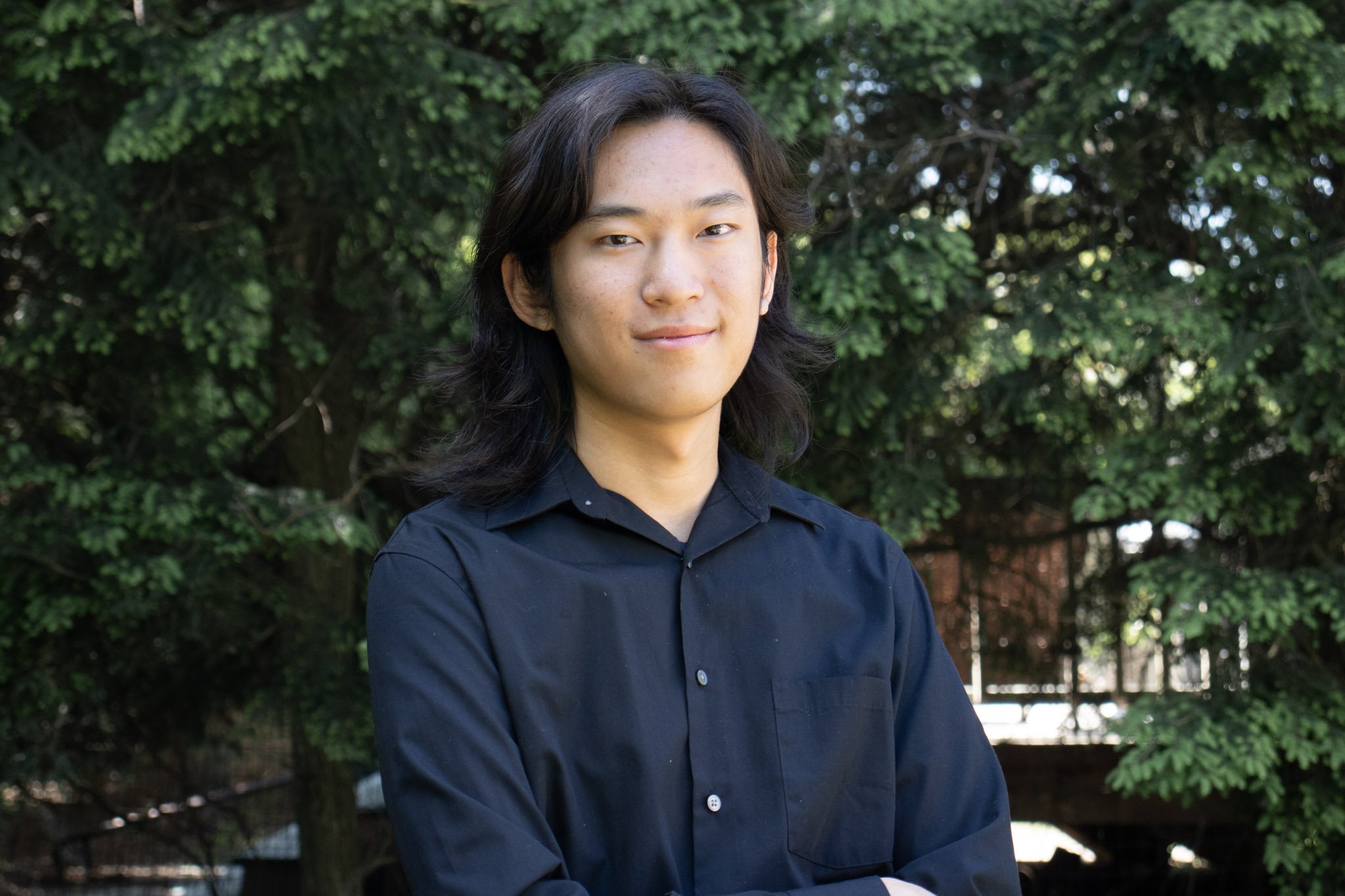 A young man with long black hair and light skin, wearing a dark blue button-up shirt, stands outdoors in front of green leaves and trees, looking at the camera with a slight smile.