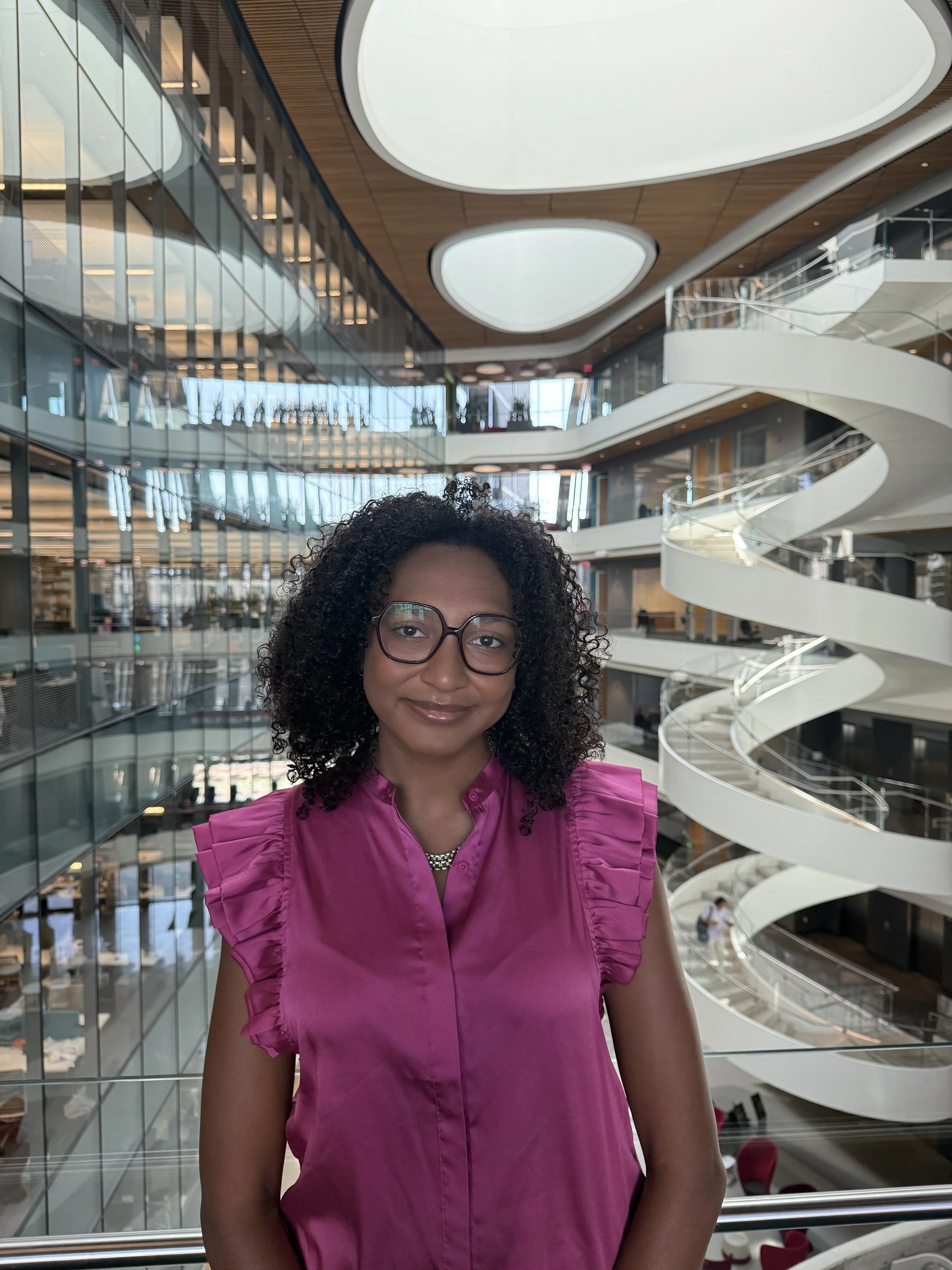 A woman with curly hair and glasses wearing a pink top, smiling inside a modern building with multiple floors and a spiral staircase.