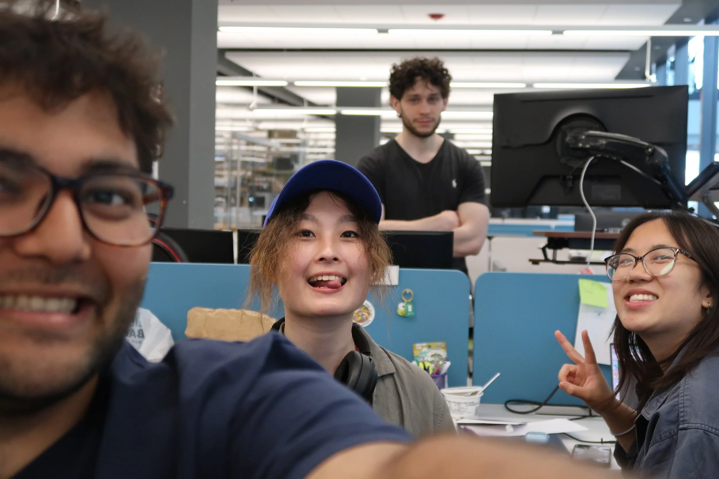 A group of people taking a selfie in an office or library setting, with a young woman wearing a blue cap making a silly face and a young woman making a peace sign, while a man with curly hair and a beard looks on in the background.