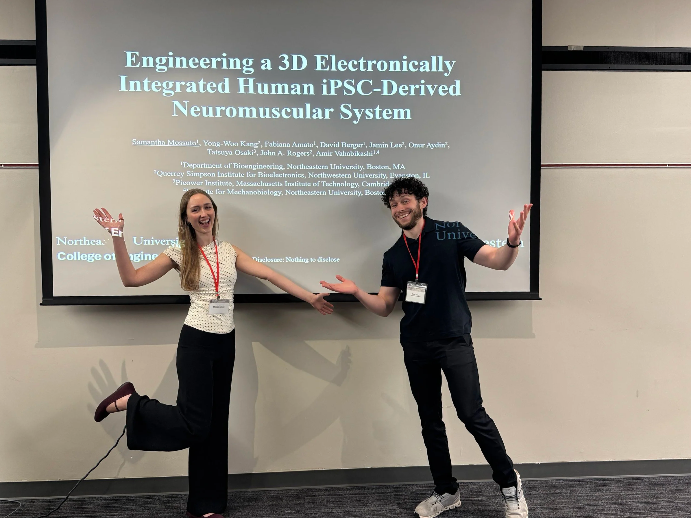 Two individuals, a woman and a man, are smiling and posing playfully in front of a presentation screen. The woman is on the left, standing on one leg with her right arm raised, and the man is on the right, doing a similar pose with his left arm raised. The screen behind them displays a research presentation titled 'Engineering a 3D Electronically Integrated Human iPSC-Derived Neuromuscular System' with author names and affiliations.
