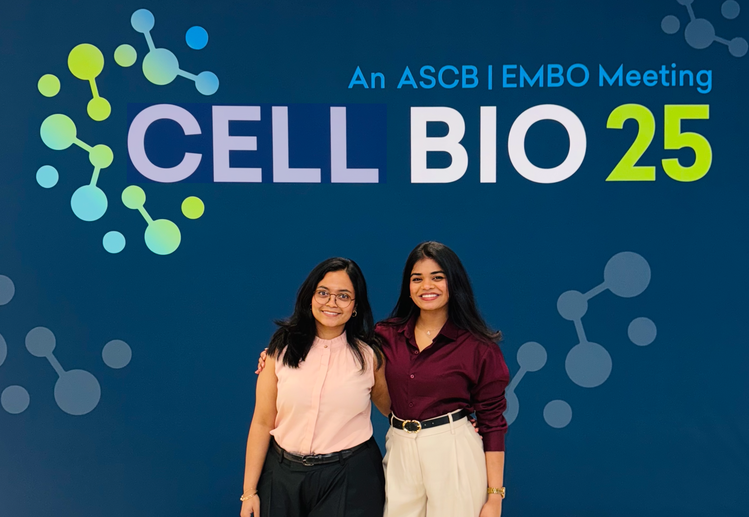Two women standing together smiling at a conference, with a blue backdrop displaying the text 'An ASCB | EMBO Meeting CELL BIO 25' and molecular graphic designs.