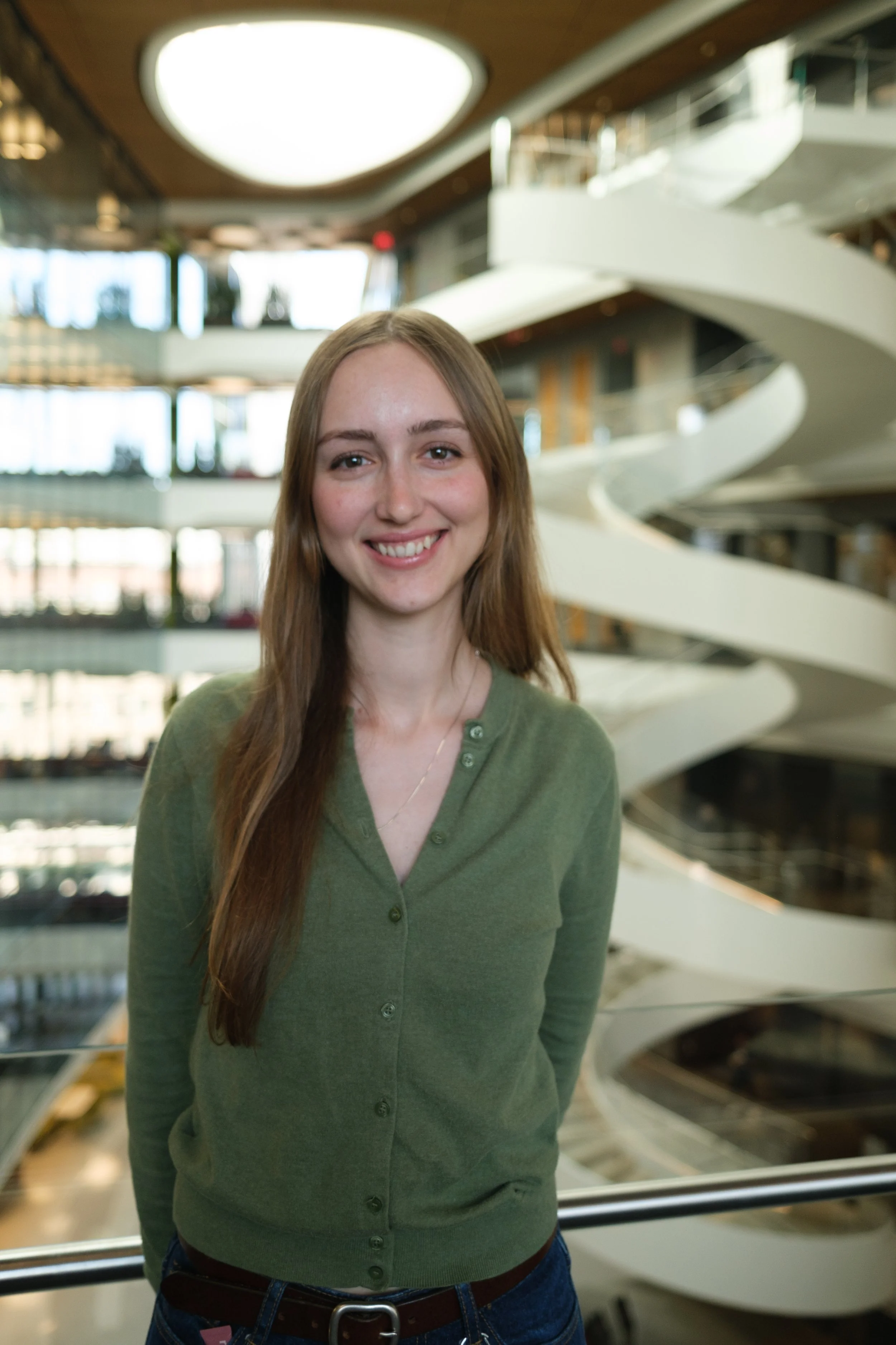 A young woman with long brown hair smiling, standing indoors with a modern spiral staircase in the background.