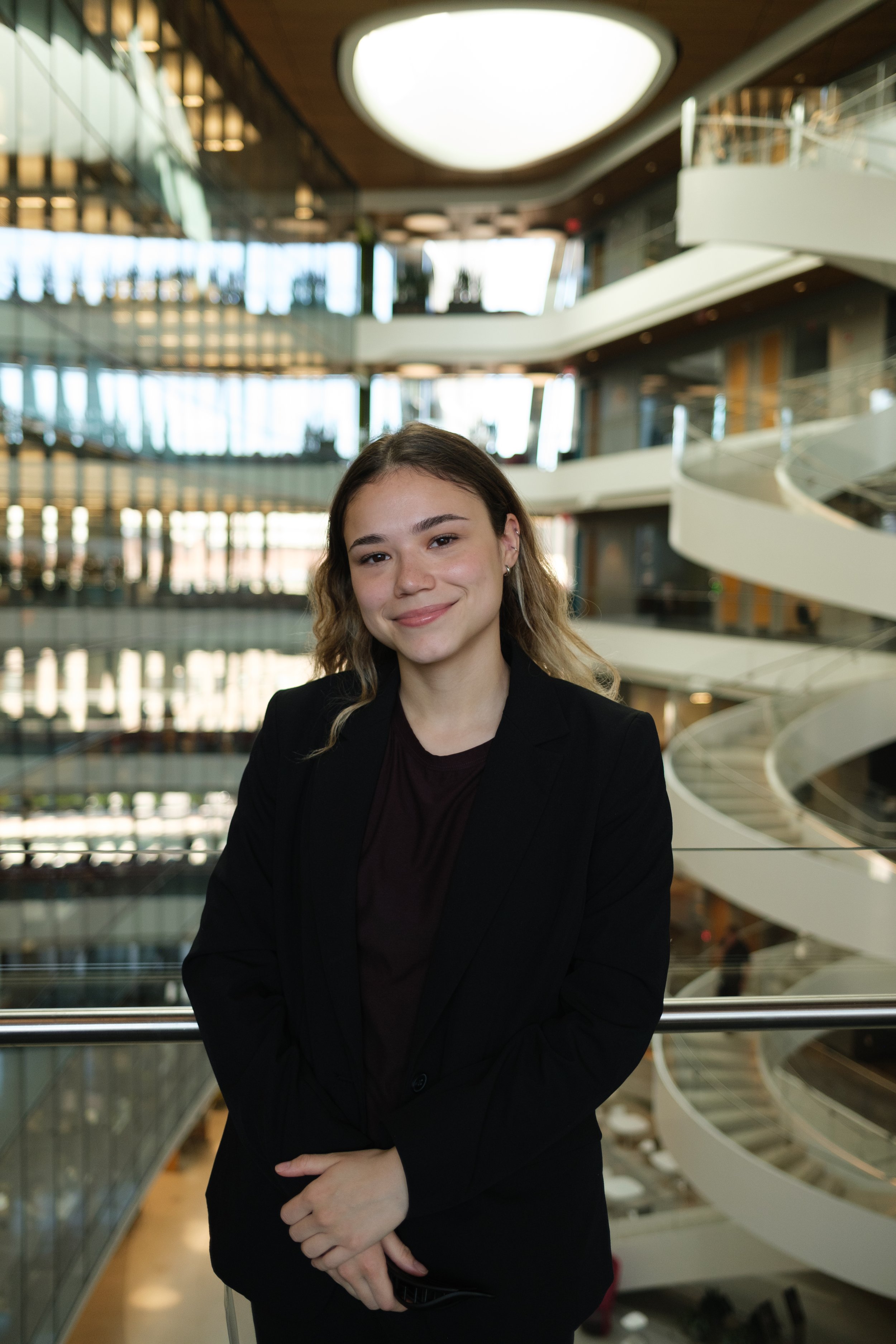 A smiling young woman with wavy hair wearing a dark blazer, standing in a modern multi-story building with glass railings and a spiral staircase behind her.