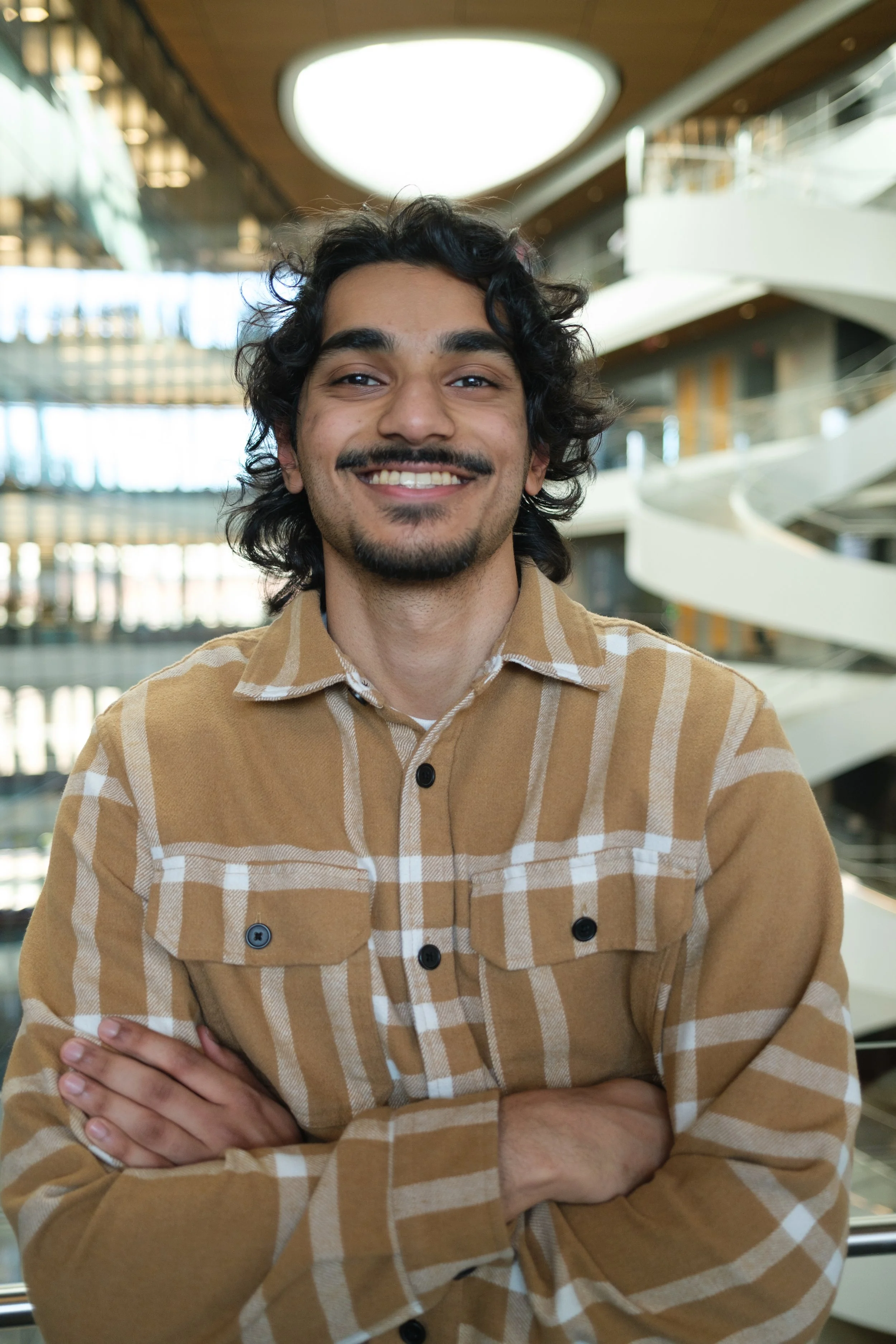 A young man with curly dark hair, a mustache, and a beard, smiling with arms crossed, wearing a beige checkered jacket, standing inside a modern building with glass and wooden elements.