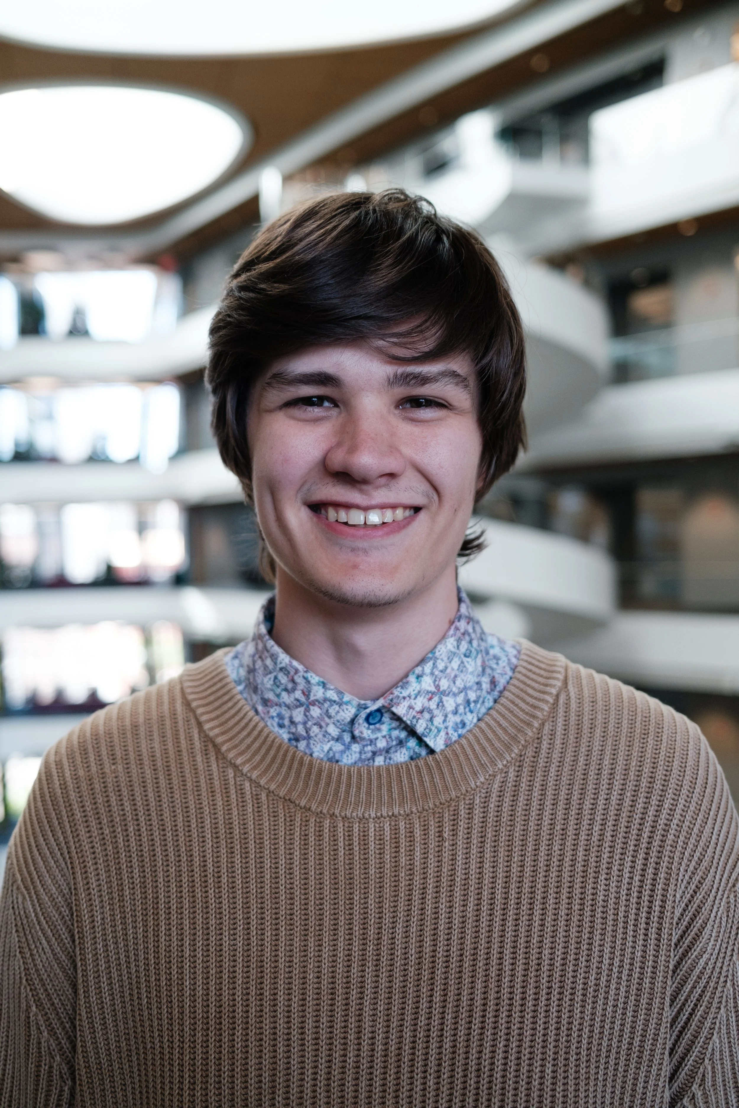 Portrait of a young man with dark brown hair, smiling, wearing a patterned shirt and a tan sweater, in an indoor space with multiple floors and natural light.