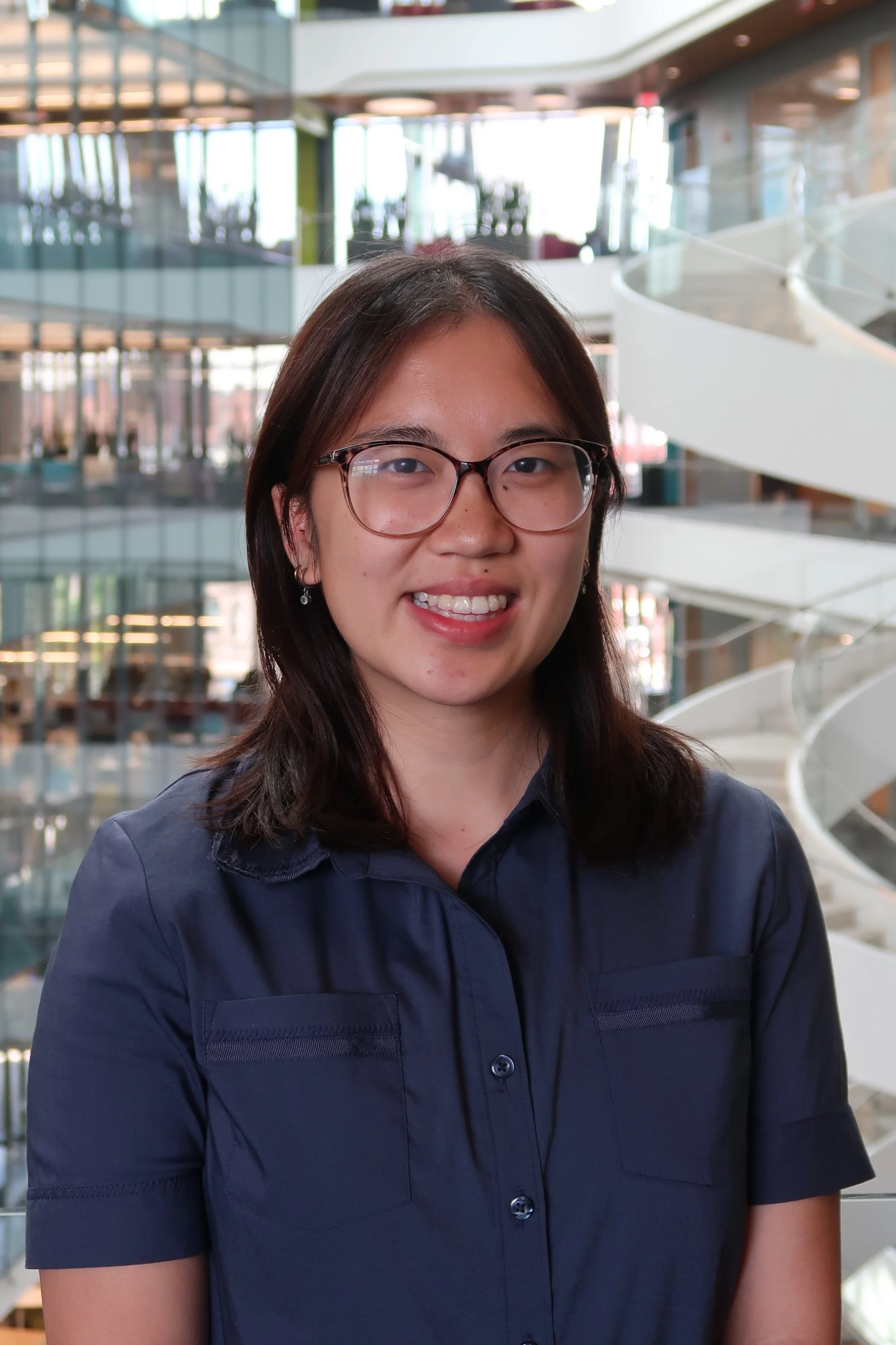 A young woman with glasses and earrings smiling, wearing a navy blue shirt, in an indoor space with modern architecture and a winding staircase in the background.