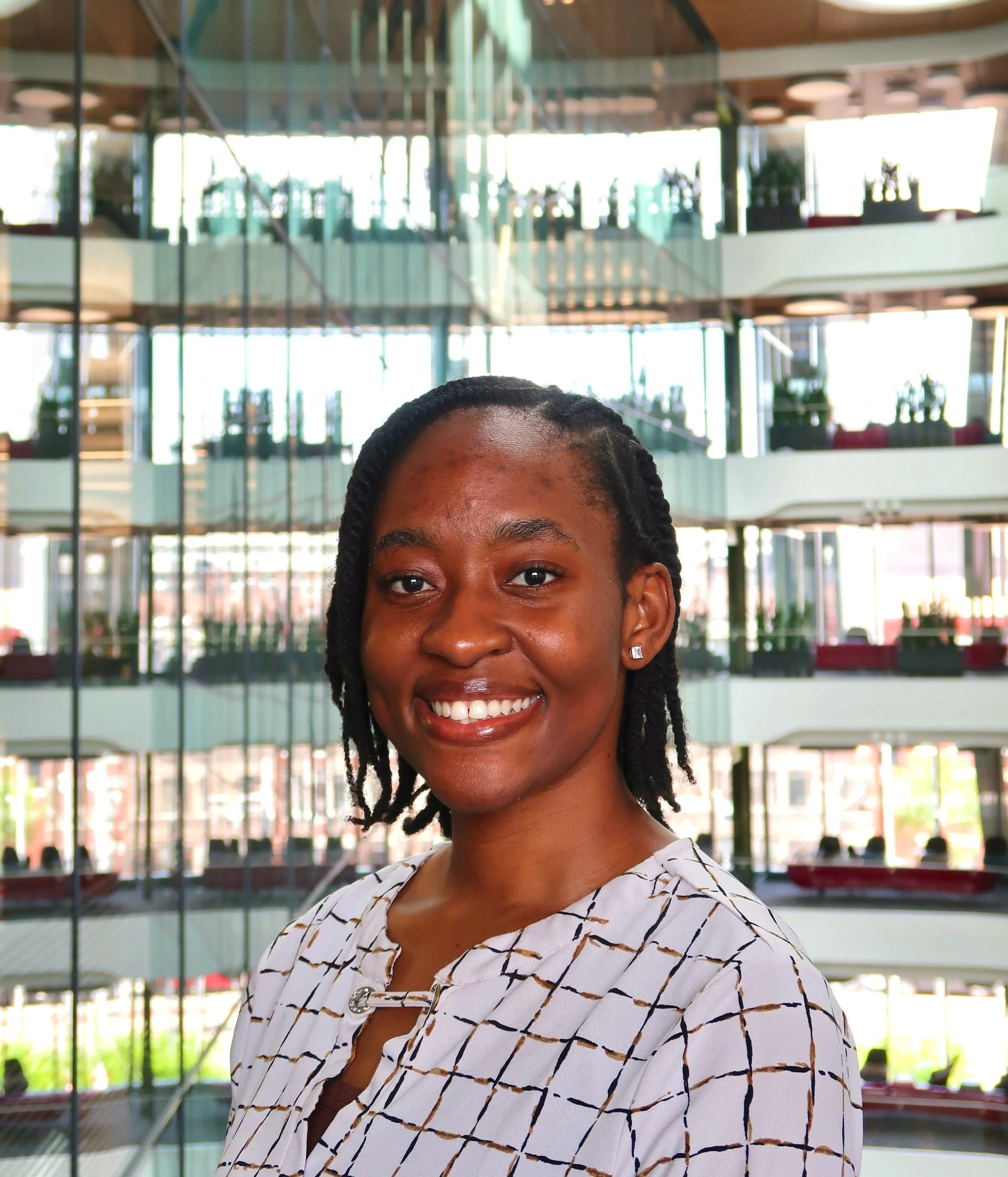 A woman with a big smile, wearing a white top with black and brown patterns and earrings, standing indoors with a background of glass walls and a multi-level building structure.