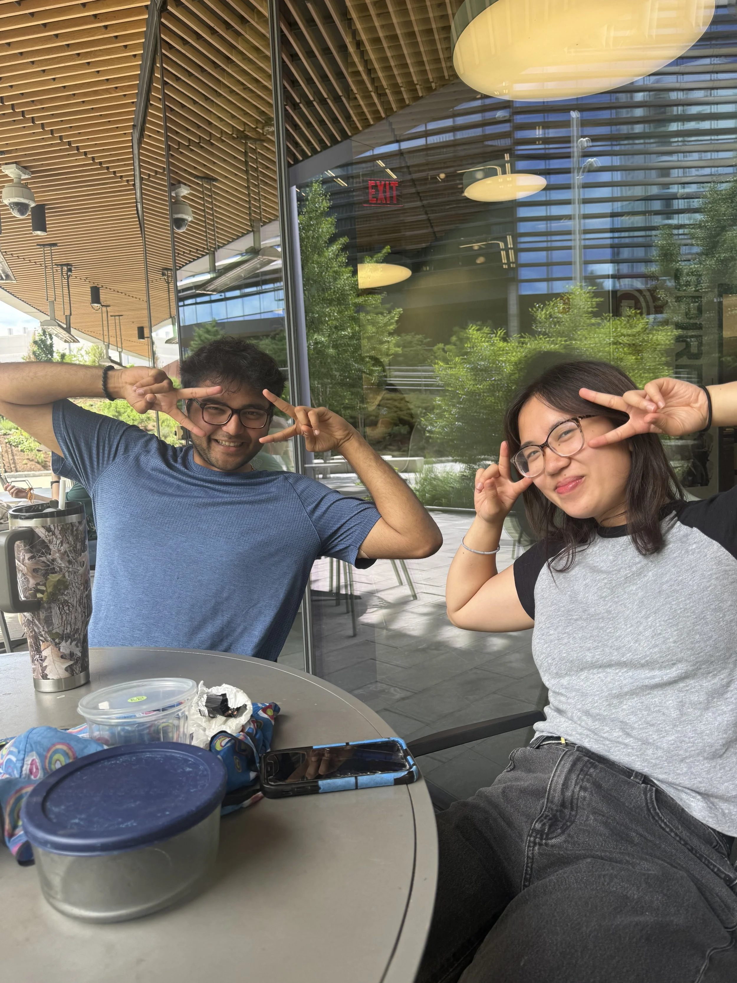 Two people, a man and a woman, sitting at a table outside a building with glass windows and a wooden ceiling, making peace signs near their faces and smiling.