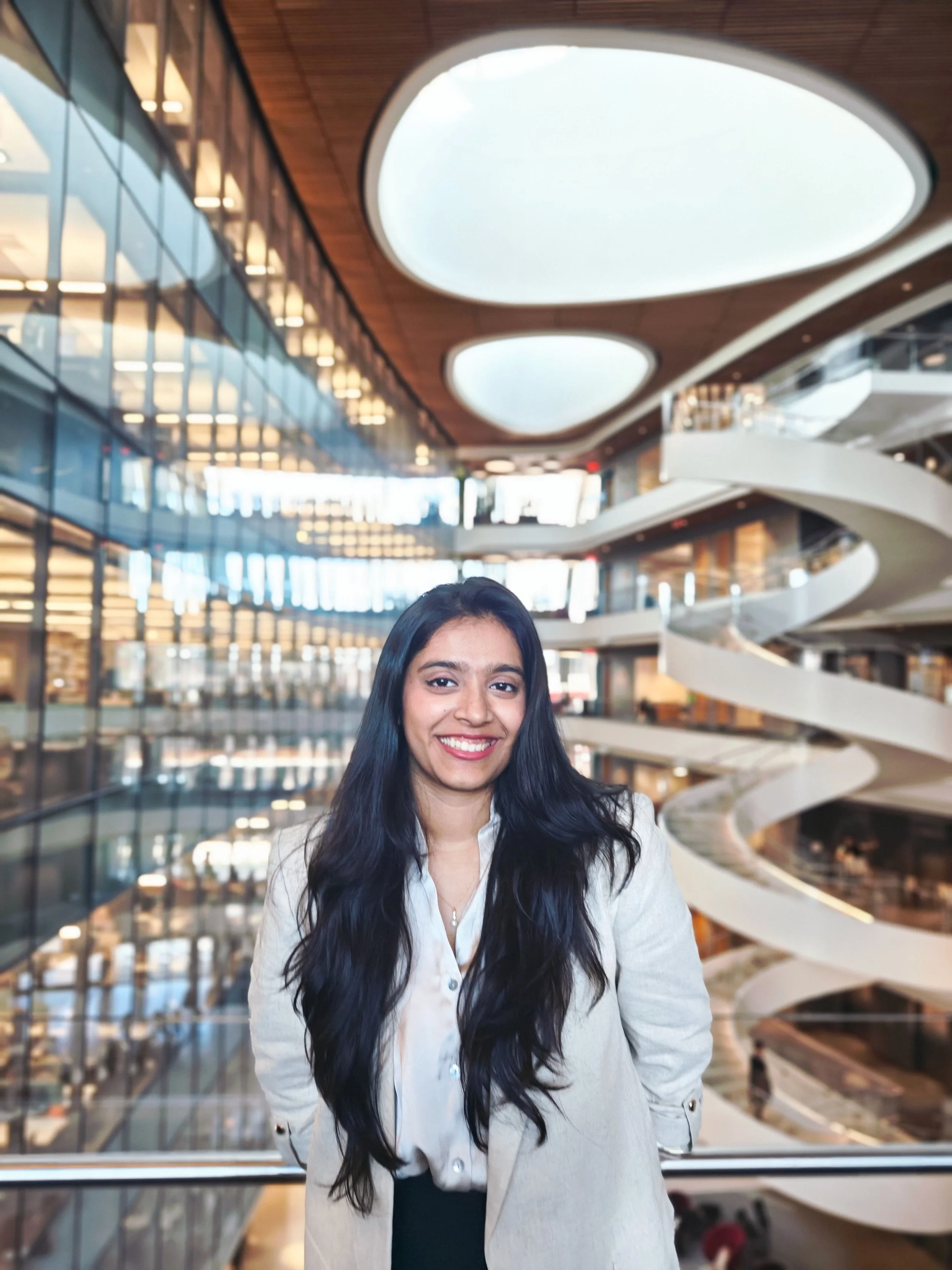 A woman with long black hair smiling, wearing a white blazer and black top, standing in a modern building with curved white balconies and a large glass window wall.